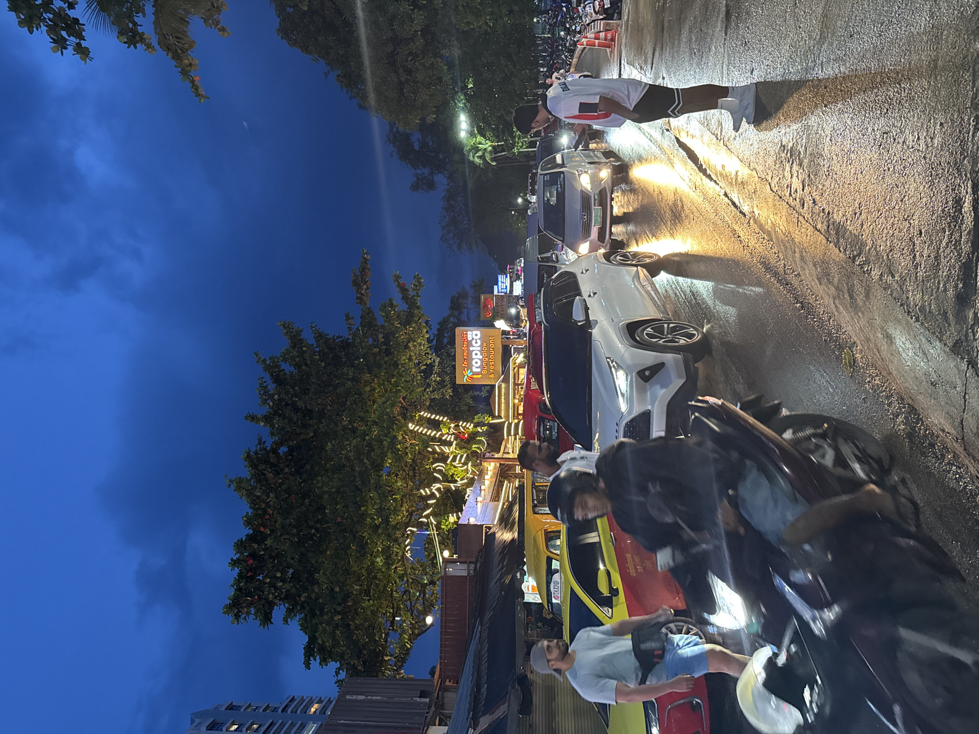 The image depicts a street scene at dusk, likely in a tropical or subtropical location, given the lush trees and building architecture. The sky is a deep blue, suggesting the transition from day to night. The ground appears wet, possibly from recent rain, and reflects the artificial lights.
The scene is populated by several people, mostly captured in motion or interacting with vehicles. One person stands on the right side of the road, illuminated by headlights, wearing a white shirt with red and black accents and black shorts. Several vehicles are visible, including cars and motorcycles, creating a sense of moderate traffic. One car, in particular, is a white SUV. A motorcycle in the foreground is dark colored.
Several signs are present. One prominent sign, lit with neon or LED lights, reads "Tropica Bungalow & restaurant". Another sign, partially obscured, displays the word "BUNGALOW". String lights are draped across some trees and structures, adding to the ambiance. The overall mood seems to be casual and lively, with people engaged in everyday activities. The time of day is evening, and the wet road suggests potential rain or high humidity. The presence of multiple vehicles and illuminated signs indicates an urban or semi-urban commercial area.