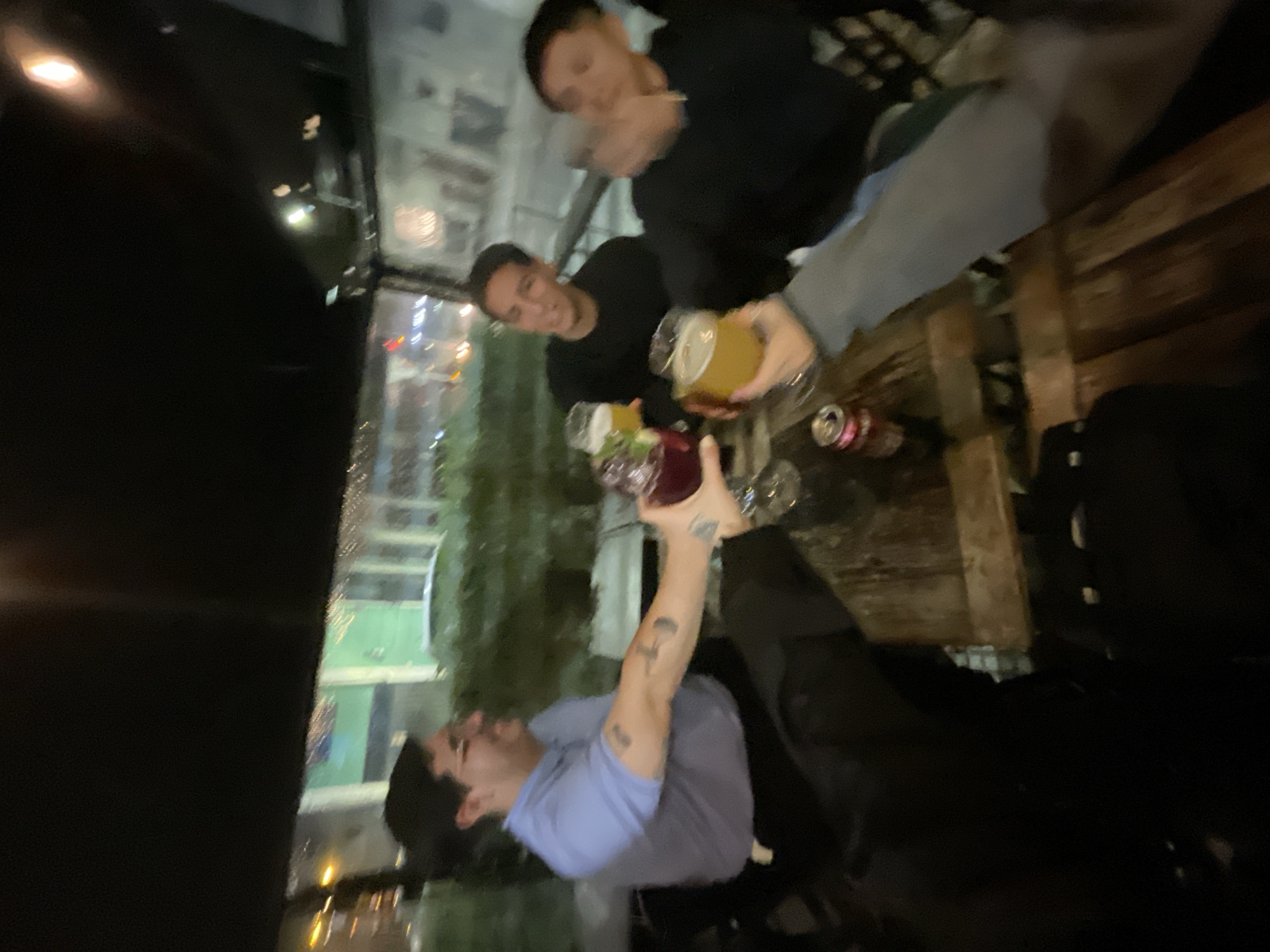 Four people are seated at a wooden table, toasting drinks. Two of the drinks appear to be beer in clear glass mugs, one is a dark red drink in a stemmed glass, and another is a mix of colors in a clear glass. Two cans are also on the table. The men are wearing casual clothing. A large window with a dark frame shows nighttime city lights and a possible park or green space. The location may be a restaurant or bar in the city of Buenos Aires, Argentina.