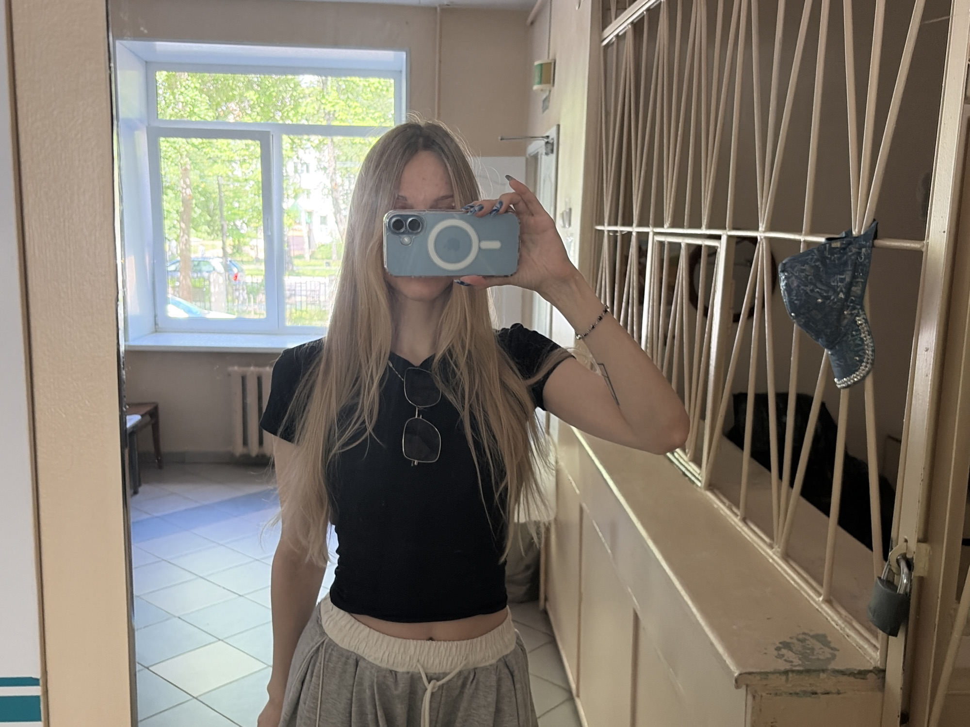 A young woman with long blonde hair is taking a selfie in a mirror. She is wearing a black crop top, grey sweatpants, and sunglasses hanging from her neck. The phone she is using to take the photo is light blue and has a circular white object on the back. She is standing in what appears to be a hallway or a communal area, possibly in an apartment building.
Behind her, a window shows trees and greenery, suggesting it is daytime and possibly spring or summer. The hallway has tiled floors and beige walls. To her right, there is a metal gate with a padlock, and a dark blue sparkly item is hanging on the gate. The overall impression is that of a casual, everyday moment.