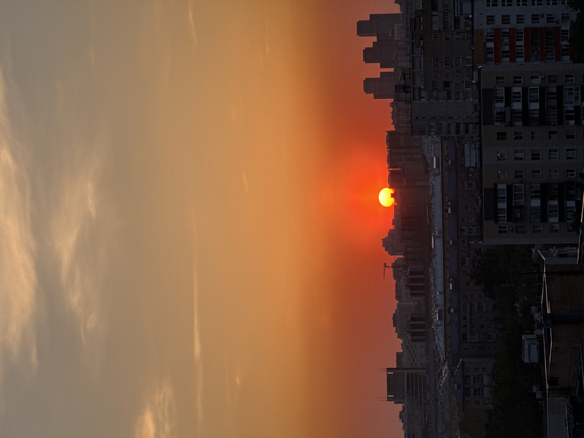 The image shows a sunset over Moscow, Russia. The sun is a bright orange orb, partially obscured by buildings on the horizon. The sky is filled with an orange and yellow gradient that fades into a darker blue near the top. The city skyline is visible as a silhouette against the bright sky. Buildings of various sizes and shapes make up the skyline, some appearing to be older apartment buildings and others newer skyscrapers. A crane can be seen on the horizon. The perspective is from a high vantage point, possibly a rooftop or high-rise building.