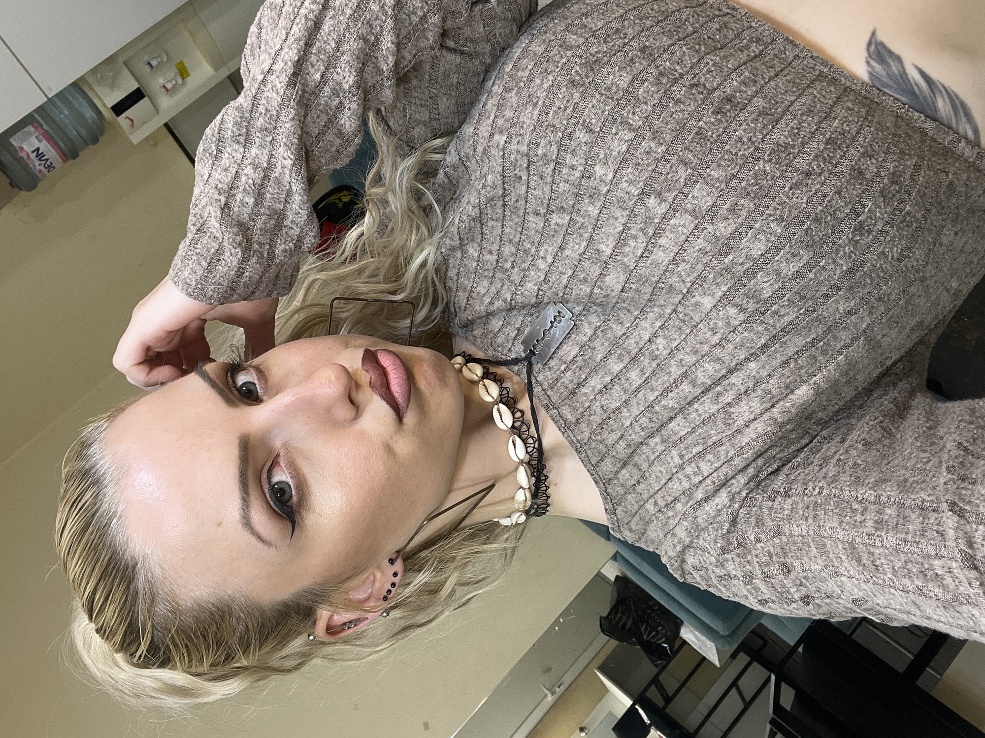 A woman with blond hair and visible ear piercings is posing indoors. She is wearing a ribbed, long-sleeved top and a necklace with shells. Her makeup is visible. In the background, there is a water bottle labeled "Devin" and a partially visible shelf or cabinet. Parts of furniture are also visible in the lower right corner. The location is unknown, not certainly Plovdiv, Bulgaria.