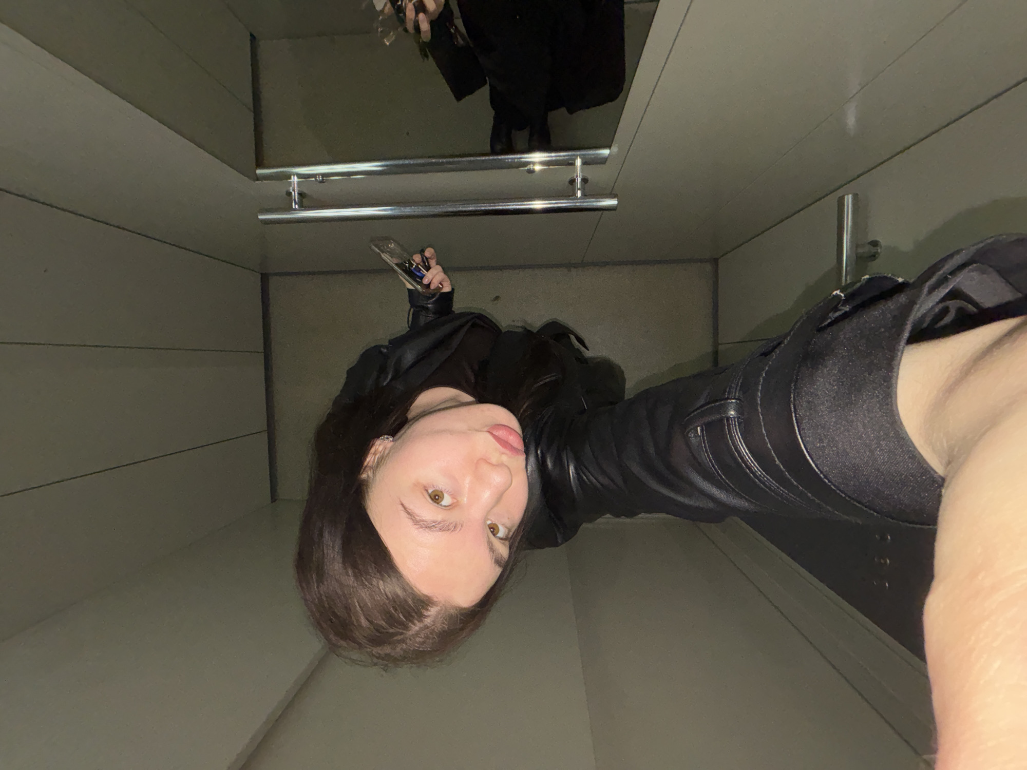 A high-angle selfie captures a young woman looking upwards towards the camera, which she holds extended in her right hand along with a set of keys. Her face, positioned in the lower-middle, shows light brown eyes and a slightly playful expression with her tongue partially out. She has medium-length dark brown hair and is wearing a black top and a black leather jacket. A small silver earring is visible in her left ear.
The setting appears to be an enclosed space, possibly a small room or cubicle, with light-colored, possibly paneled walls. In the upper portion of the image, two horizontal chrome-colored bars are prominent, reflecting light. Above these bars, a highly reflective surface, likely a mirror, shows the inverted reflection of a person's lower body, including dark pants and shoes. The lighting is even and appears to be indoor artificial light. No distinct text is visible in the image.