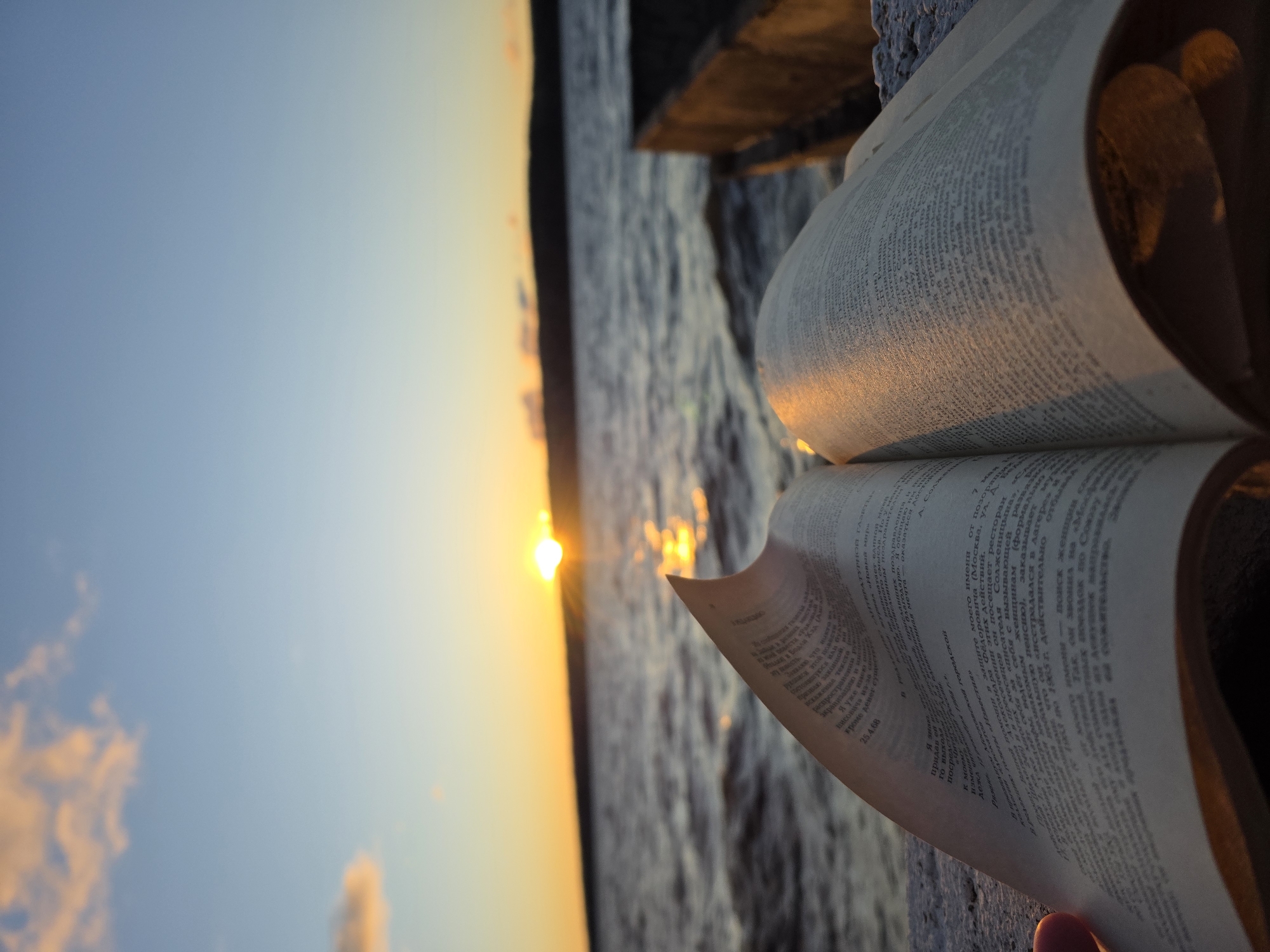 The image depicts an open book with Russian text overlaid against a blurry background featuring a sunset over water. The book appears to be resting on a concrete ledge. The location is implied to be Chaikovsky, Russia. The text visible on the book's pages includes phrases such as "Раковый", "Москва" and "городской". There are no people visible in the scene. The overall environment is one of quiet contemplation, with the sunset providing a warm glow on the book.