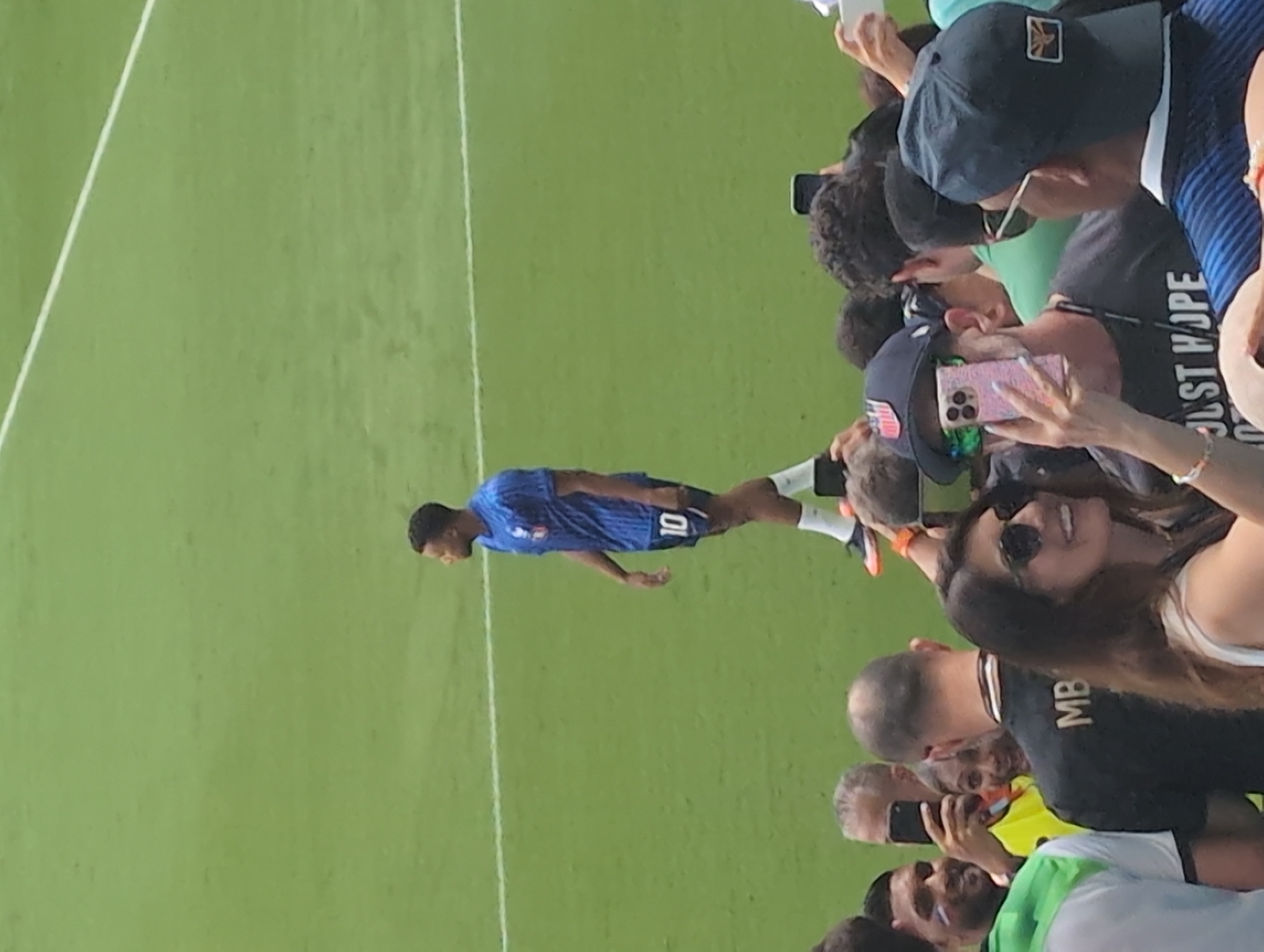 This is a medium shot, eye-level, outdoor photograph taken at a soccer stadium in Miami Gardens, Florida, United States of America. The image captures a crowd of spectators gathered behind a soccer player who is standing on a green field. The player is wearing a blue soccer jersey with the number 10 on the back and black shorts. He is looking to his right, away from the camera. The spectators in the foreground are mostly out of focus, but some are holding up their phones, presumably to take pictures of the player. A woman in the foreground is wearing sunglasses and a white top, and she is smiling and holding up her phone. The field is marked with white lines, and the background is a blur of green grass.