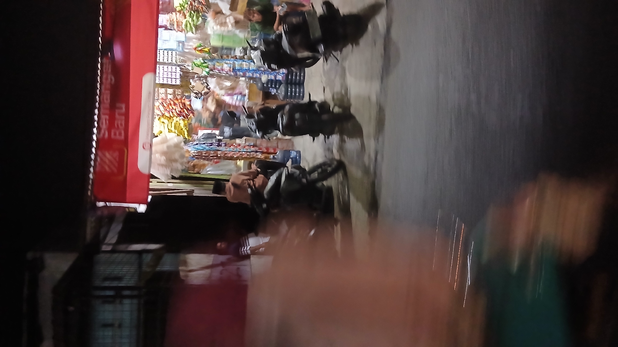 The image depicts a street scene at night in Klambir Lima Kebun, Indonesia. A red awning with white text that reads "Semangat Baru" is visible above a small store. The store appears to be selling various goods, including packaged snacks and drinks, with some items displayed on shelves and hanging from the ceiling. Several motorcycles are parked in front of the store, and at least three people are visible: one person sitting on a motorcycle, another sitting on a stool near the store entrance, and a third person sitting on a bench in front of the store. The lighting suggests artificial illumination from the store and possibly streetlights. The foreground is blurred, indicating motion blur, possibly from the camera operator or a passing vehicle.