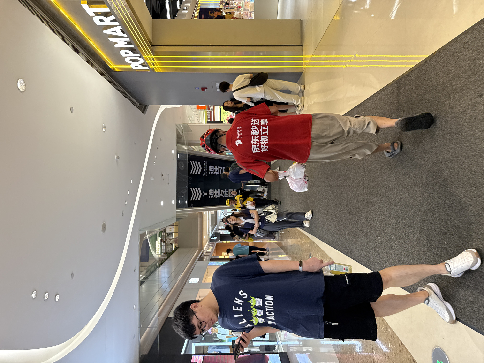 This is a vertical, full-body shot of people walking in a shopping mall. The setting appears to be a modern, well-lit indoor space with a mix of tiled and carpeted flooring. In the foreground, a person wearing a dark blue t-shirt with "LIENS ACTION" and cartoon aliens printed on it is looking down at their phone. They are wearing black shorts and white sneakers.
Behind this person, a delivery person in a red t-shirt with Chinese text that appears to translate to "JD.com Express, Enjoy Good Things" and a red helmet is walking away from the camera. They are carrying a white plastic bag. To the left, a prominent "POP MART" sign is visible, illuminated by yellow lights, indicating a retail store. Other shoppers are seen in the background, some walking and others standing. The lighting suggests it is daytime. The overall atmosphere is casual and everyday.
