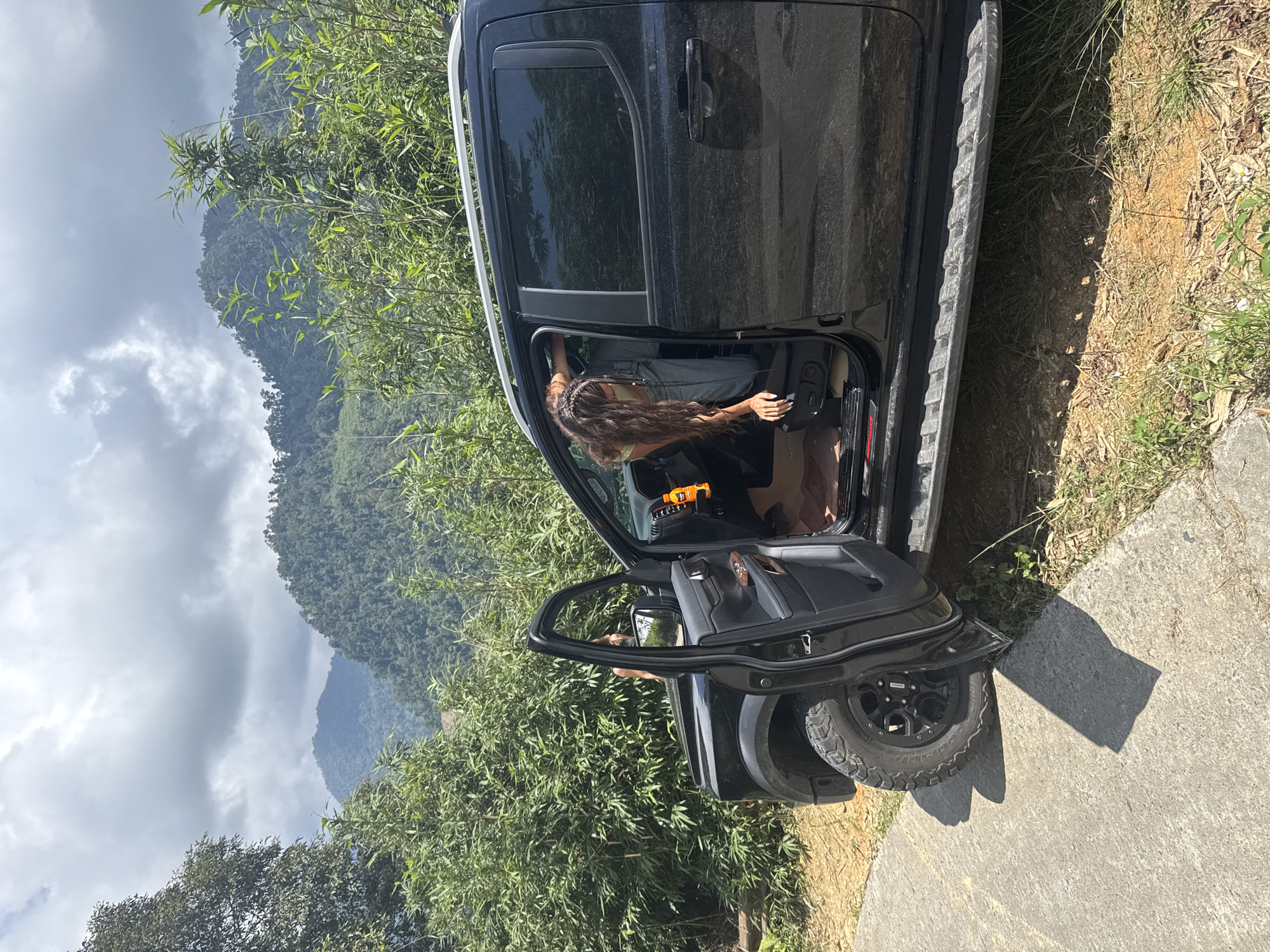 A black pickup truck is parked on a concrete road in what appears to be a mountainous, rural environment. The driver's side door is open and a person with long, dark hair is visible inside the vehicle, partially obscured by the door frame. The surrounding environment includes green vegetation, including bamboo, and distant, cloud-covered mountains. Based on the description, the location is Suối Giàng, Vietnam.