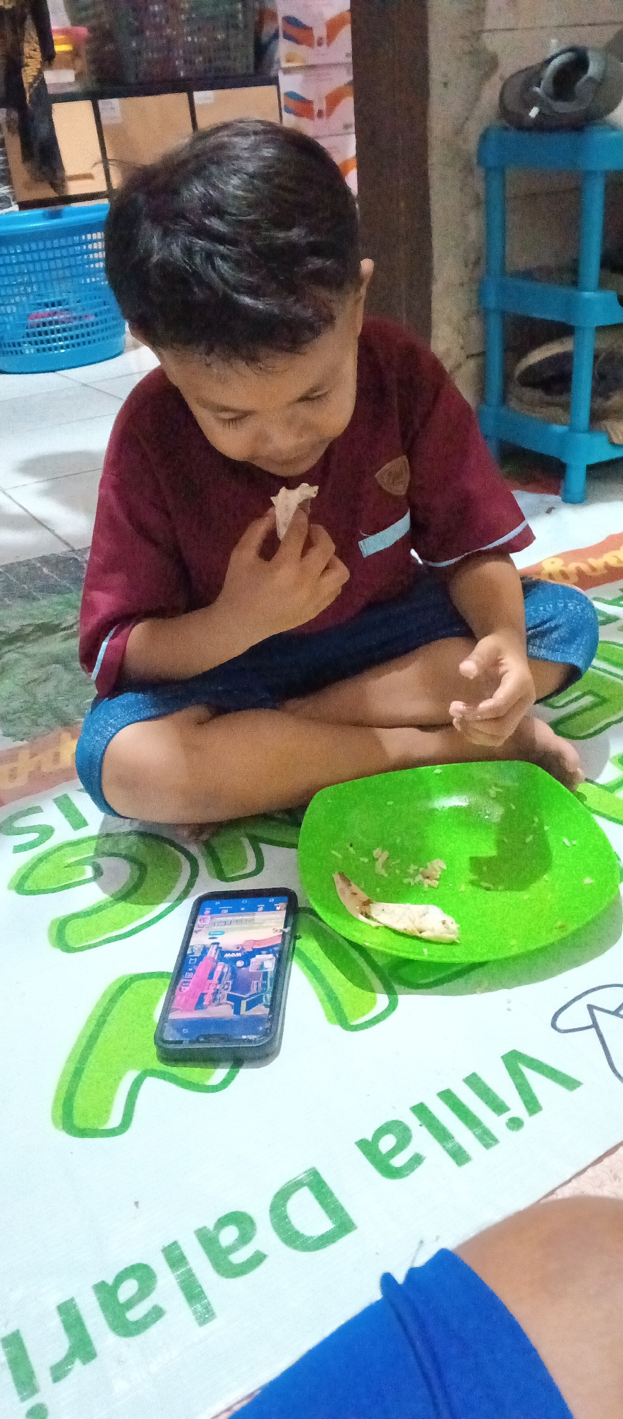 A young Indonesian boy is sitting cross-legged on a mat with text that appears to read "Villa Dalari" and some other unreadable text. He is wearing a maroon shirt and blue shorts. In his right hand, he is holding a small piece of food up to his mouth and appears to be eating it. To his right is a green plastic plate with some leftover food. In front of him is a smartphone showing a colorful screen, possibly a game or app. The background shows a blue plastic basket and some shelves with various items. The scene is indoors, with a tiled floor.