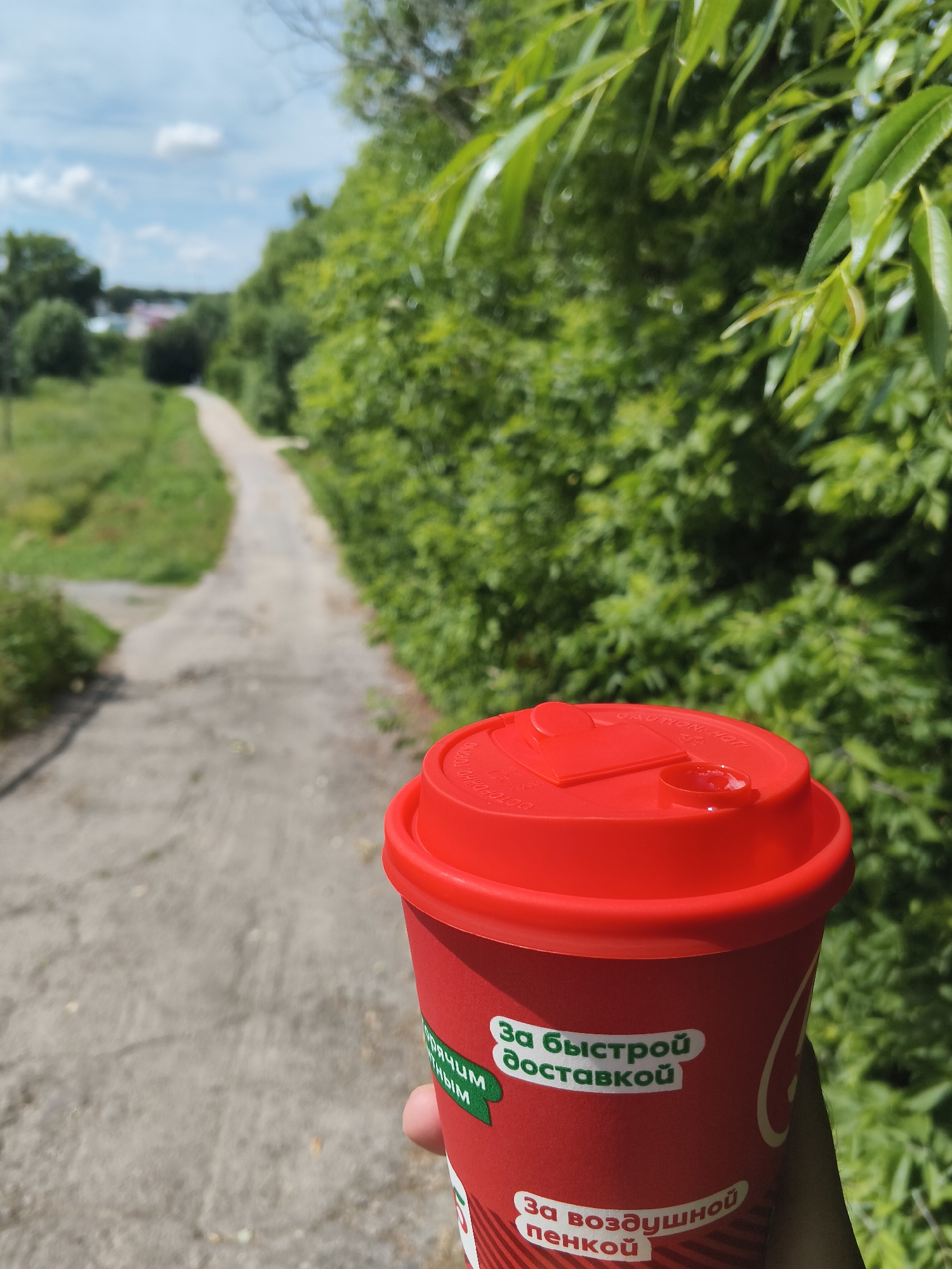 In Mokshan, Russia, a hand holds a red paper coffee cup with a red lid. The cup has text in Russian, including "За быстрой доставкой" (For fast delivery) and "За воздушной пенкой" (For airy foam). The background is a blurred scene of a dirt path leading through a grassy area with trees and green foliage under a cloudy sky. The perspective is from the viewpoint of someone holding the cup, looking down a path.