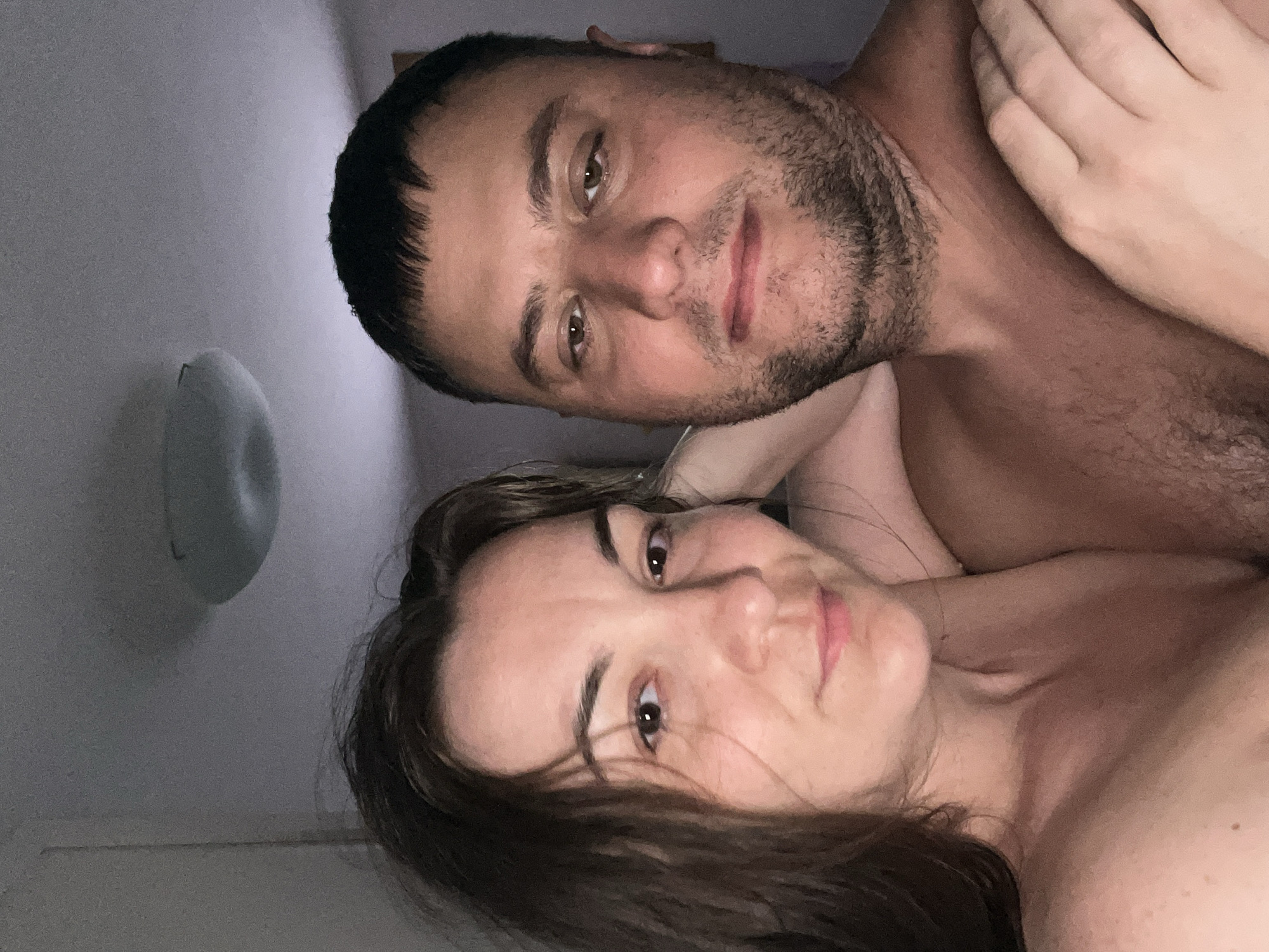 A medium shot selfie captures a man and a woman from the chest up, positioned closely together. The man, on the right, has short, dark hair and a stubbled beard. His head is tilted slightly forward, and his eyes are open. The woman, on the left, has long, dark brown hair that falls around her face. Her head is tilted to the side, and she is looking towards the camera with her eyes open. Both individuals appear to be shirtless. They are indoors, with a ceiling light fixture visible in the background. The room's walls are painted a neutral color. The overall lighting is dim, suggesting it may be evening or nighttime. There are no discernible landmarks or features that would indicate the specific location in Acquaviva, Italy.