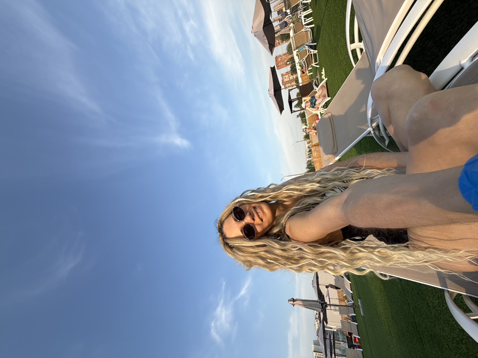 The image shows a woman with long, blonde dreadlocks and sunglasses lying on a white chaise lounge chair. She is smiling and appears to be taking a selfie. The background features a clear blue sky with wispy clouds. Other chaise lounge chairs with umbrellas can be seen in the background on what appears to be artificial turf. In the distance, there are buildings and trees. This scene could be located in the city of Шебунино, Russia, although the specific location cannot be determined from this image.