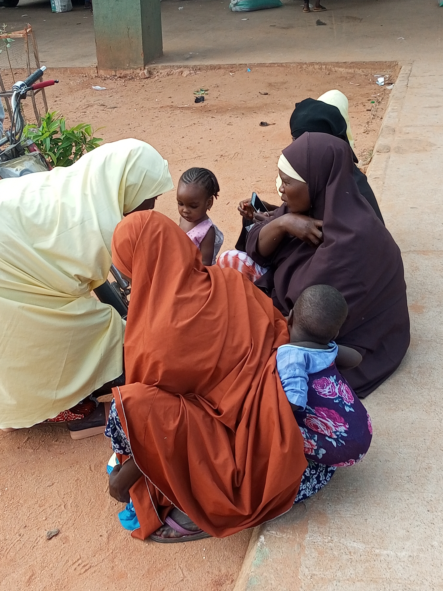 This image captures a moment of everyday life in Gombe, Nigeria, likely near a marketplace or public gathering area. The scene is set on a dirt and paved surface, with a building and possibly market stalls in the background. A bicycle is parked on the left side of the frame.
The central focus is a group of at least four individuals, three women and two young children, gathered together. One woman, wearing a light yellow hijab, is partially visible from the back. Another woman, in a dark brown hijab, is holding a mobile phone and appears to be interacting with it. A third woman, wearing a bright orange hijab, is crouched down, with her back to the camera. Two young children are also present. One child, a girl with braided hair, is wearing a pink top and is looking downwards. Another child, a boy in a blue shirt with a floral pattern, is seated and facing away from the camera.
The individuals are dressed in traditional Islamic attire, with headscarves and long garments. Their postures suggest a casual gathering, perhaps resting or socializing. The environment appears somewhat dusty and worn, consistent with an outdoor public space in Nigeria. The lighting suggests it is daytime.