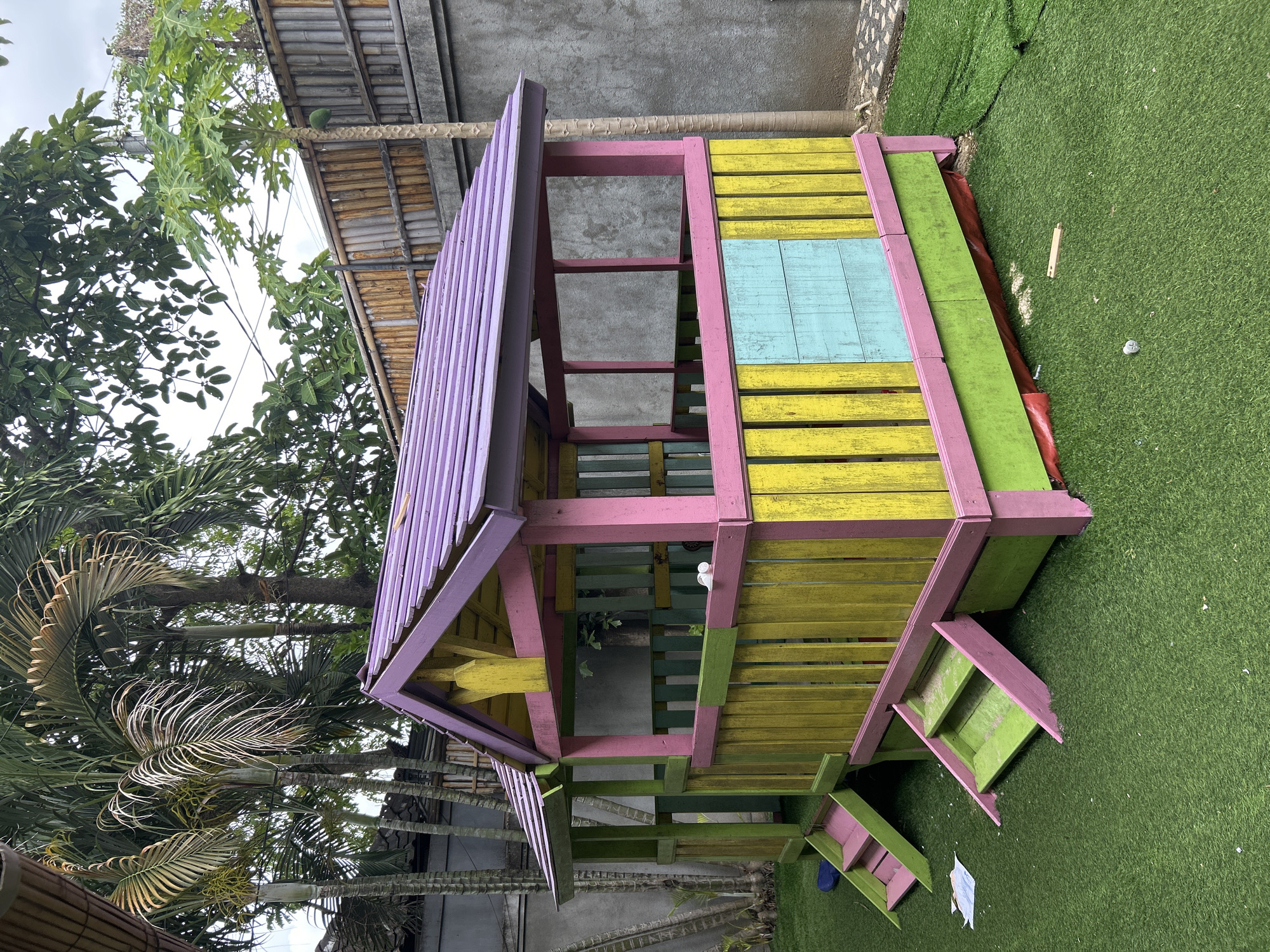 A brightly colored wooden playhouse sits on artificial turf in Kuta, Indonesia. The playhouse features a purple corrugated roof, pink and yellow trim, and a fence painted with alternating yellow and green vertical planks. A light blue panel is inset into the fence on the right side. Two sets of stairs, one pink and green and the other green with pink trim, lead up to the playhouse. In the background, palm trees and a bamboo fence are visible, with a concrete wall behind them. The sky is overcast.