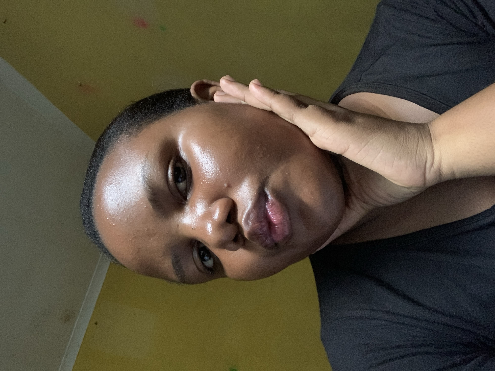 A close-up, angled shot of a woman with dark skin, possibly indoors. She's wearing a black top. Her left hand is touching her face, near her ear and cheek. The background is a pale yellow wall. Given the request for location specifics, it's not possible to ascertain if this photo was taken in Dar es-Salaam, Tanzania, without any further information. The photo's content provides no clear connection to the city or country mentioned.