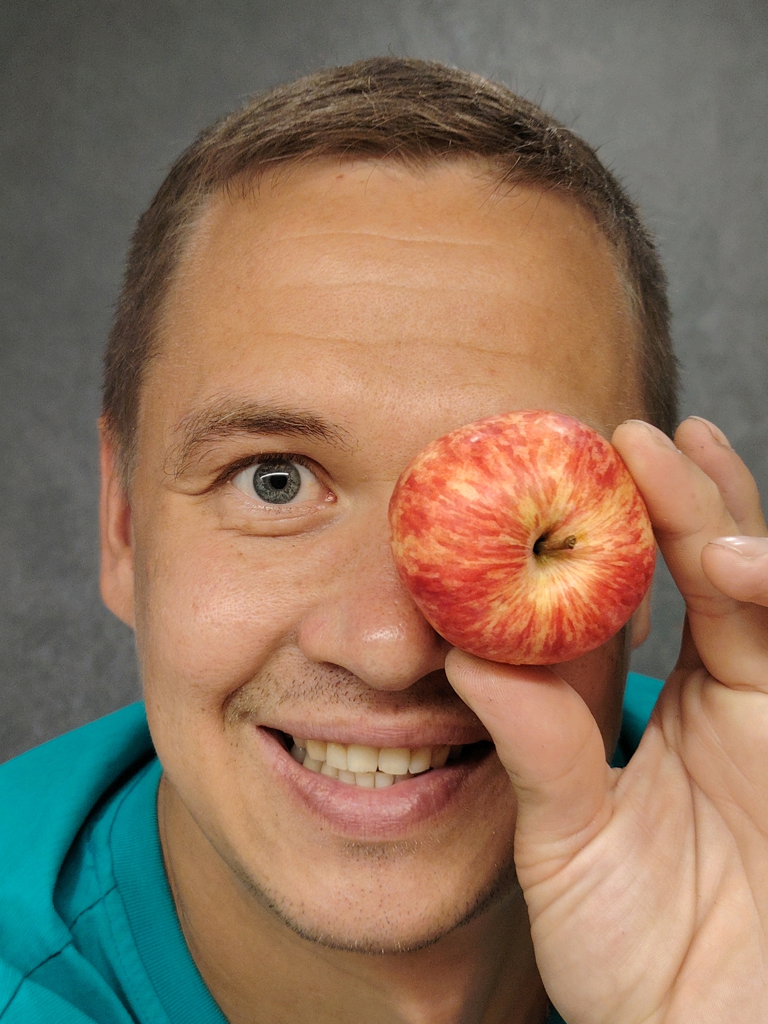 A man's face is shown from the chest up, with a gray, textured background. He has short, brown hair and is wearing a teal shirt. His right eye is visible, and he is holding a red and yellow apple in front of his left eye with his right hand. His mouth is open in a smile, revealing his teeth. The man's skin is fair, with some stubble on his chin. The lighting is bright, casting shadows on his face. There is no discernible indication of the city of Chaikovsky, Russia, or any other specific location.