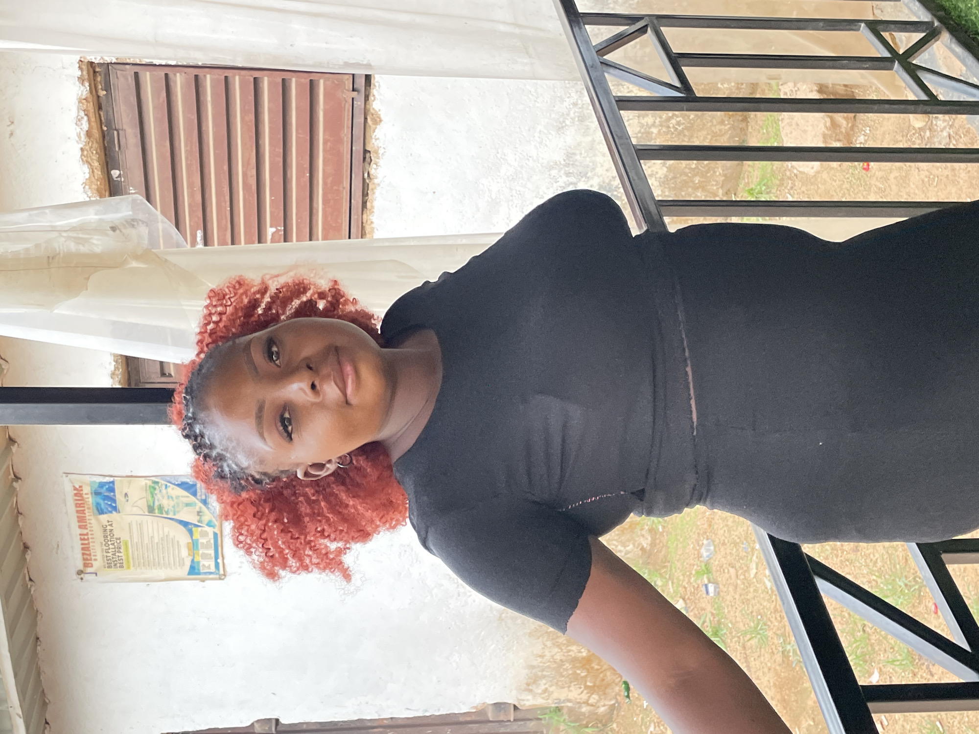 A young, dark-skinned woman with reddish-brown curly hair poses for a portrait in Barakin Akawo, Nigeria. She wears a black, form-fitting outfit. The scene is outdoors with a metal railing behind her and a weathered white wall to her left. A poster with the name "BEZALEL AMARIAK" and flooring installation information hangs on the wall. Above her, there is a small window with a metal grille. The ground visible through the railing appears to be dry with sparse vegetation.