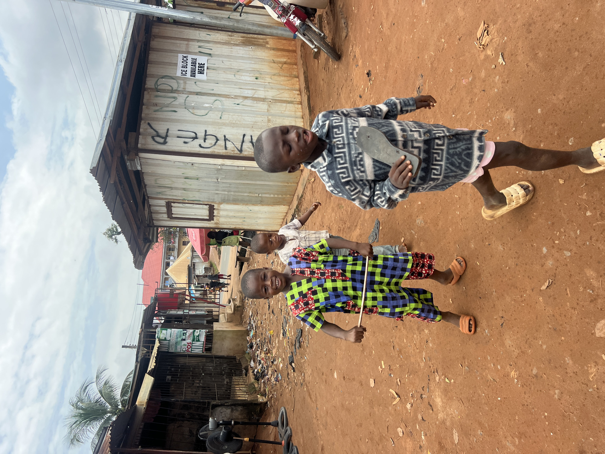 The image depicts three young boys in Nkpor, Nigeria, standing on a dirt road in front of makeshift structures. The boy in the foreground, on the left, wears a vibrant, checkered jumpsuit and smiles at the camera while holding a wooden stick. To his right, a second boy in a light-colored shirt walks behind him. A third boy, on the right, faces away from the camera and carries a shoe sole in his hand. The background features corrugated metal buildings, some with signs, and a parked motorcycle. The ground is uneven and scattered with debris, suggesting an unpaved urban environment. The sky is cloudy.