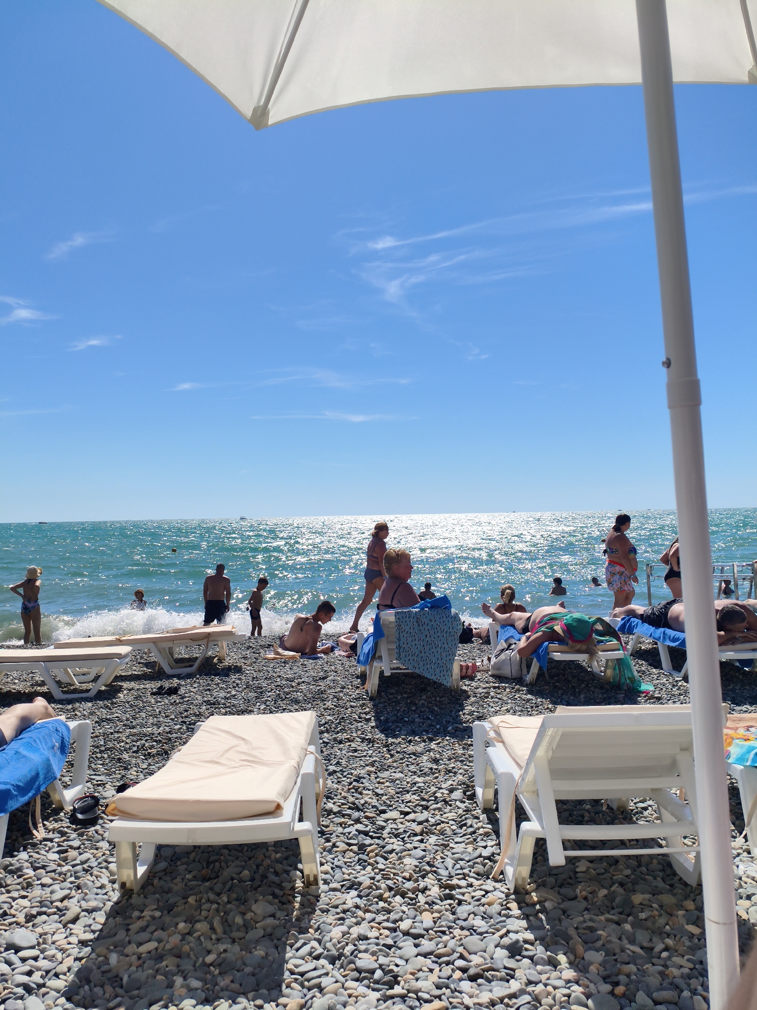 The image depicts a sunny day at a pebble beach, likely in Sochi, Russia, with people relaxing and swimming in the sea. In the foreground, several white beach loungers are set up on the stony shore. Some are occupied by individuals sunbathing, while others are empty.
A large white umbrella shades a portion of the foreground, with its pole prominently visible on the right side of the image.
The middle ground shows a group of people enjoying the water. Some are wading in the shallow waves, while others appear to be swimming further out. The sea is a vibrant blue, reflecting the bright sunlight and creating a shimmering effect.
In the background, the horizon is a clear, light blue sky with a few wispy clouds. The overall scene conveys a relaxed and leisurely atmosphere characteristic of a summer beach day.