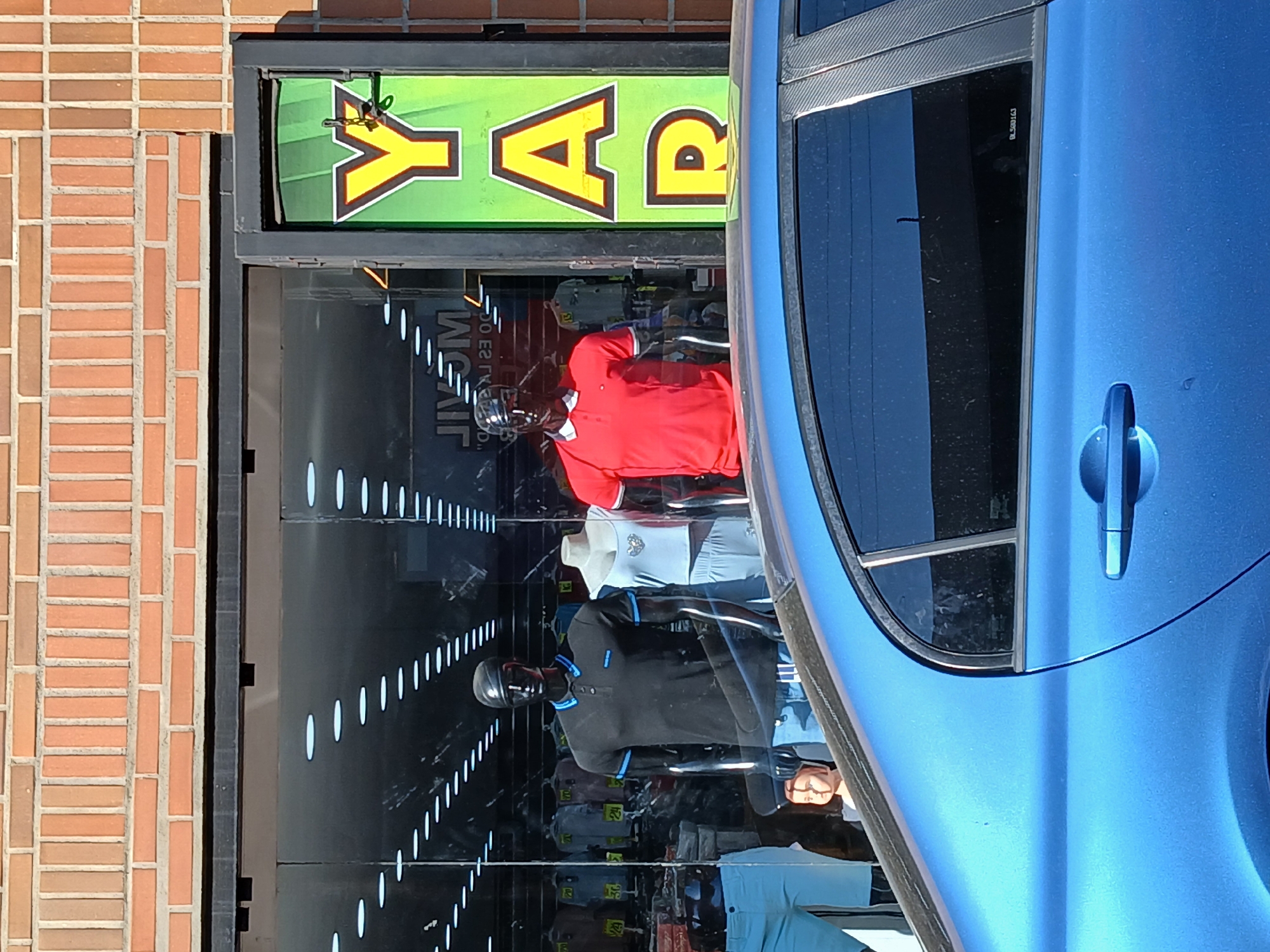 The image shows the exterior of a retail store. A portion of a blue car is visible in the foreground, reflecting the store in its windows. The store has a glass storefront, showcasing mannequins dressed in polo shirts. A sign above the door reads "RAYA" in yellow letters with a black outline on a green background. The building is constructed of brick. The location is likely Ciudad Guayana, Venezuela.
