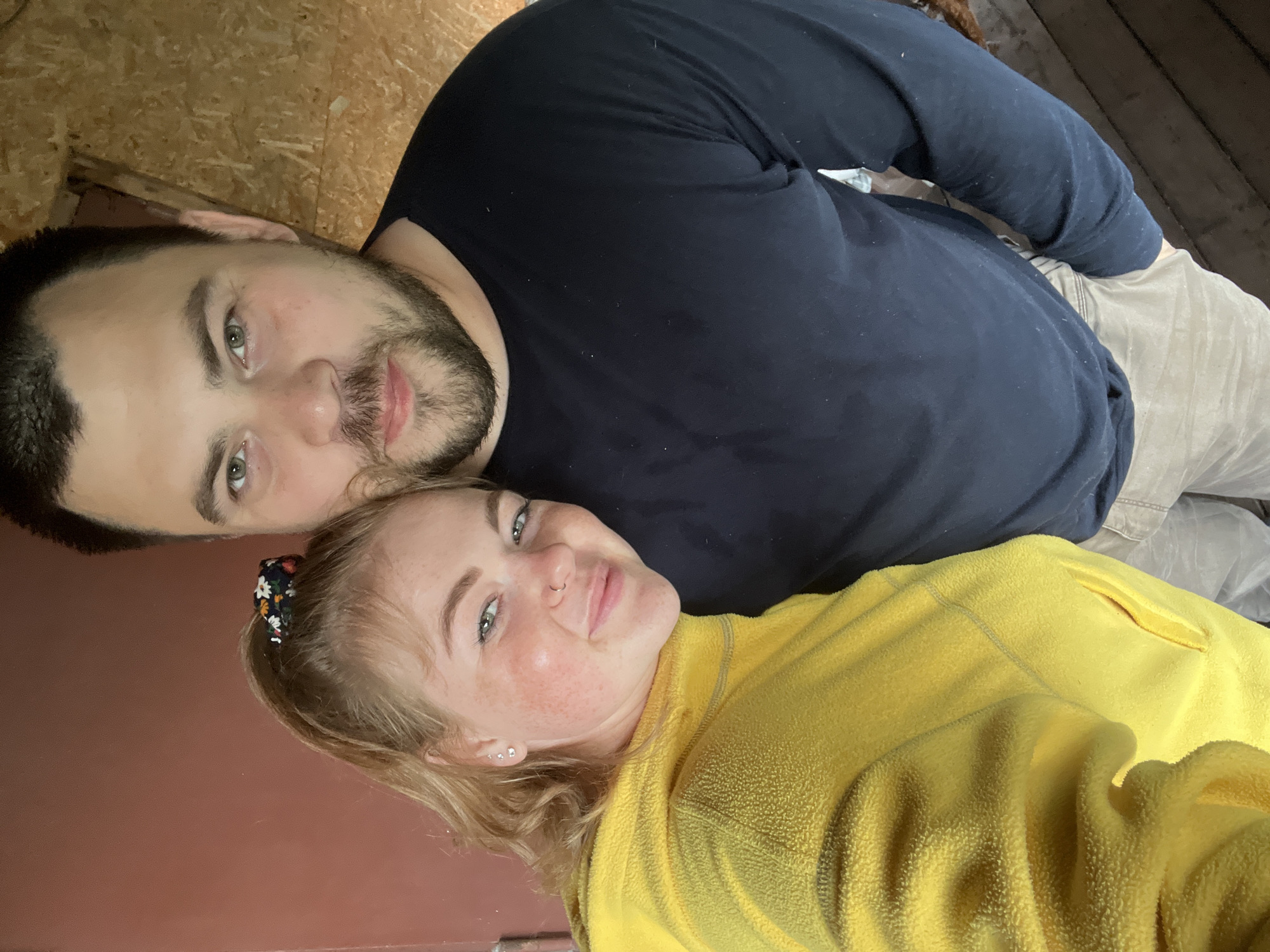This is a portrait-oriented image featuring two individuals, a man and a woman, captured from a slightly overhead perspective while they are reclining. The man, positioned on the left, has dark hair, a beard, and a mustache, with light-colored eyes. He is wearing a dark blue long-sleeved shirt and light-colored pants, and displays a subtle, content expression.
Beside him on the right is a woman with blonde hair, which is tied back with a floral-patterned scrunchie. She has visible freckles, light-colored eyes, a nose hoop, and multiple ear piercings. She is wearing a bright yellow fleece jacket and is smiling gently. Both subjects appear relaxed and comfortable.
The background suggests an indoor setting. Behind the man's head and upper body, there is a light-colored, textured surface, possibly particle board or light wood. Behind the woman and extending behind the man's lower body, there is a smooth, reddish-brown surface, likely a wall or floor. A darker wooden surface is visible in the upper right portion of the frame. The lighting is soft and diffused. No visible text is present in the image. The location context provided for this image is Chaikovsky, Russia.