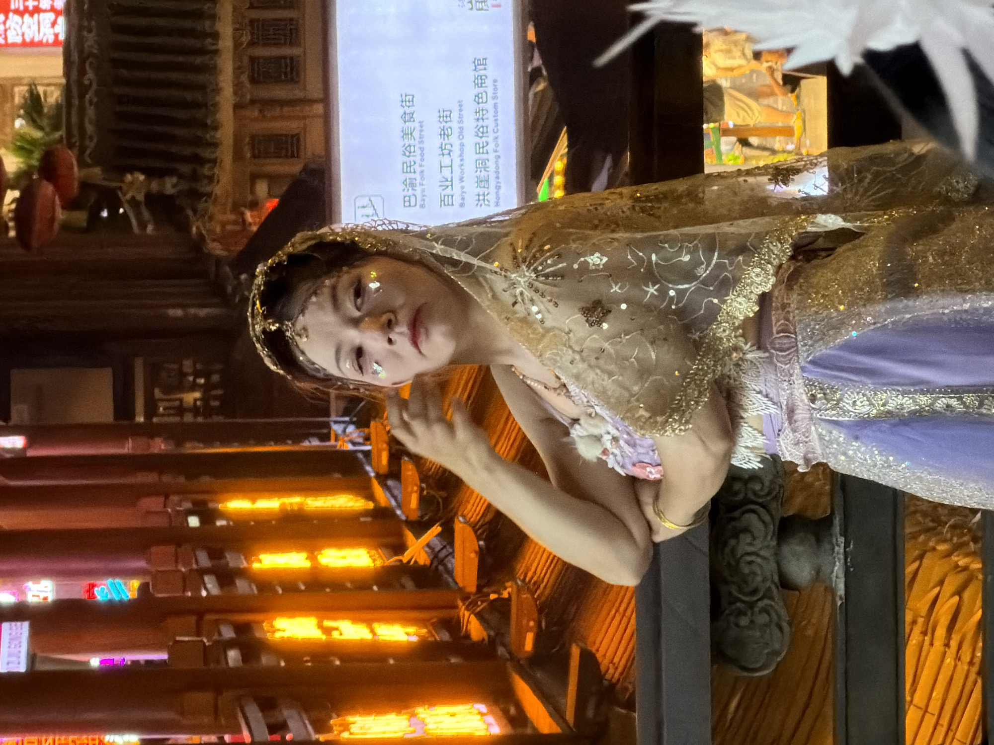 A woman in traditional Chinese attire, adorned with glitter, stands on a balcony in the Hongyadong scenic area in Yuzhong District, China. The outfit features a sheer, embellished veil and a flowing purple skirt with intricate silver patterns. She is positioned to the left of the frame, with her arm bent and hand near her face, as if posing. The background displays rows of illuminated wooden pillars and railings, typical of the architecture in this historic district. A digital screen is visible to the right, displaying Chinese text and English translations, likely indicating points of interest or street names like "Baiyu Folk Food Street" and "Hongyadong Folk Custom Store." The lighting in the scene is a mix of warm orange from the illuminated structures and cooler, artificial light from the screen and surrounding signs, creating a vibrant and somewhat atmospheric setting.