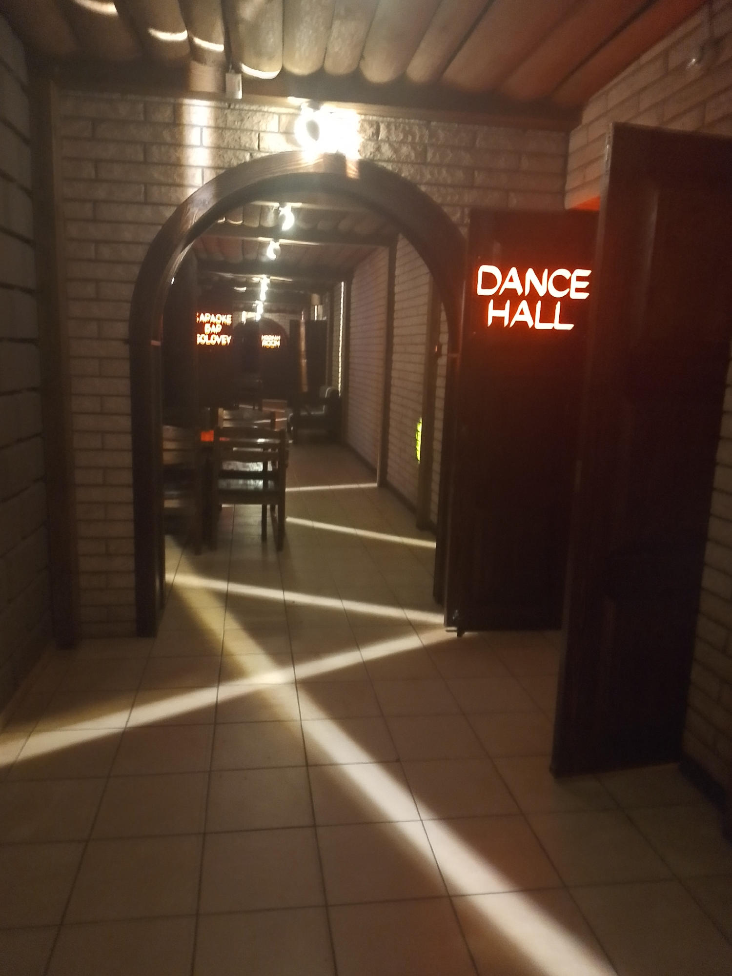 The image depicts the entrance to a "DANCE HALL" in Chaikovsky, Russia. The entrance is marked by an arched doorway constructed of brick. Beyond this doorway is a room labeled "KAPAKE BAR SOLOVIEY", which appears to be a bar with chairs and tables. The entrance to the dance hall itself is flanked by dark doors. The floor is covered in square tiles, and sunlight streams through the entrances, casting shadows across the floor. The overall lighting is dim. No people are visible in the scene.