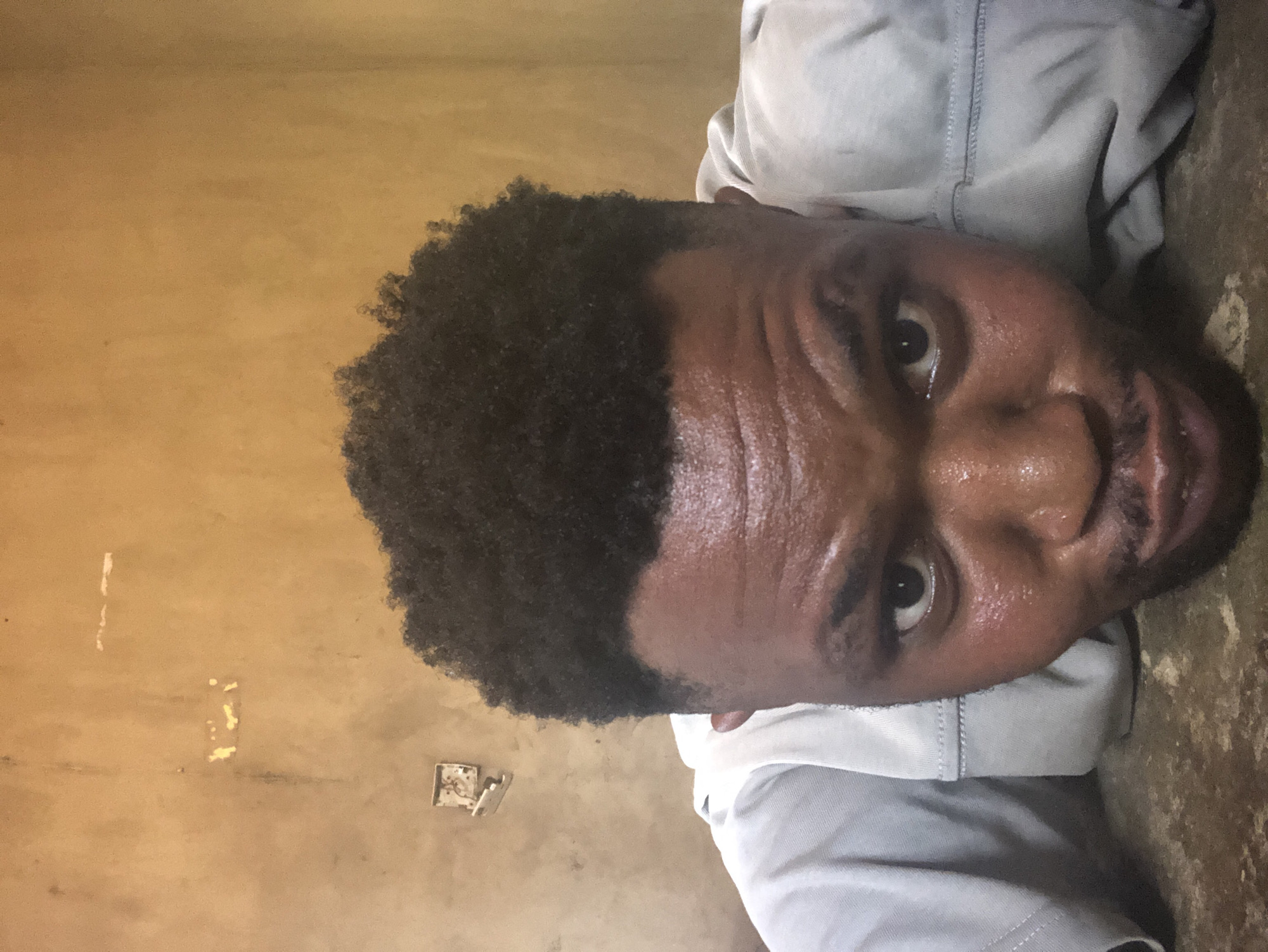 The image shows a close-up of a man's face and upper body, rotated to the side. He has dark curly hair and is wearing a light-colored shirt. His skin appears slightly shiny, possibly from sweat. He is looking directly at the camera with a slightly concerned or tired expression. The background consists of a tan wall with some imperfections and a partially visible electrical outlet. The location appears to be an indoor space, the city and specific location are unknown.