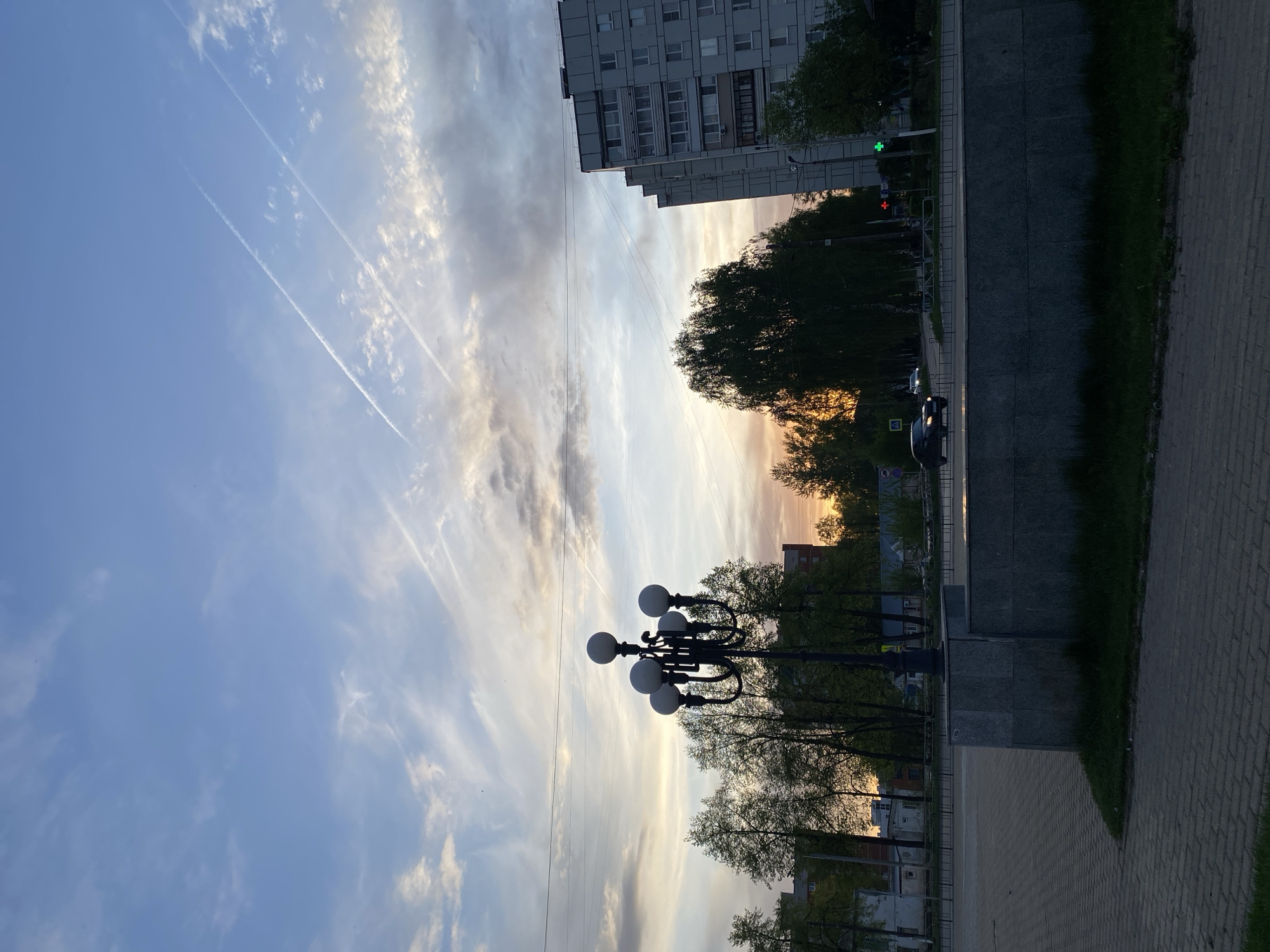 The image shows a view along a street in what appears to be the city of Chaikovsky, Russia. The sky is partly cloudy with contrails. A multi-story building is visible on the right side. A street light is positioned in the middle of the image. Trees line the street. A paved sidewalk is visible, and a street and possibly some cars are present, though the lighting makes details difficult to discern. A traffic signal shows a green light. The image appears to be taken at dusk or dawn based on the lighting.