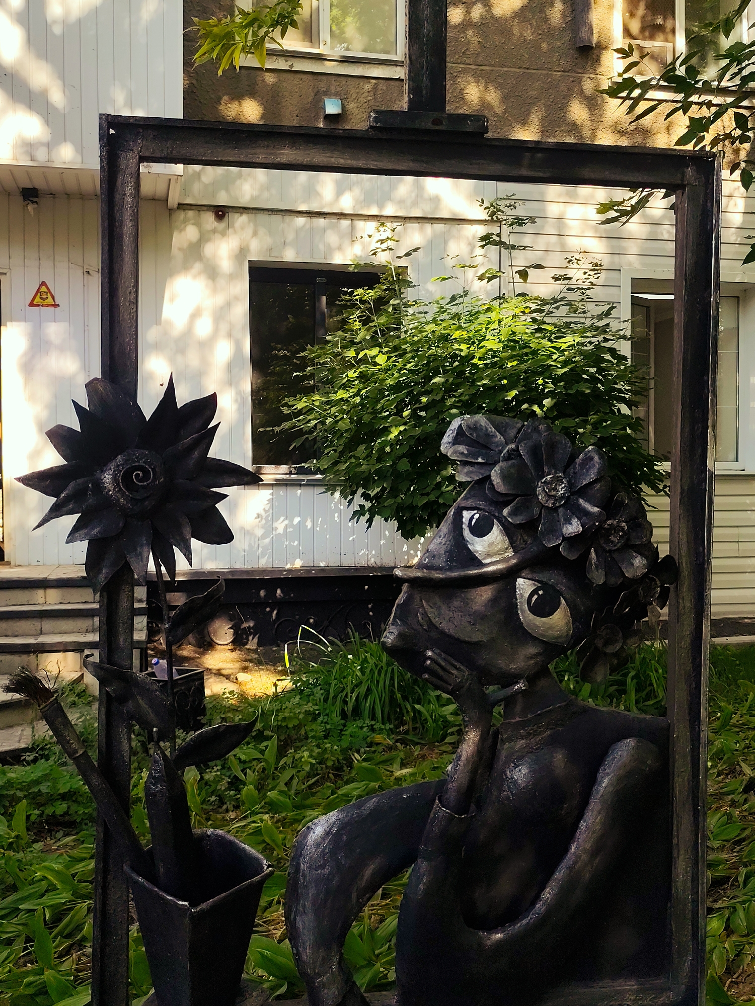 An outdoor, eye-level, medium shot captures a metal sculpture framed by a dark, rectangular metal structure. The sculpture depicts a stylized, dark figure with large, round eyes, adorned with metal flowers in its hair. It is seated with one hand resting near its mouth and a subtle smile. To the left of the figure, a large, dark metal sunflower is mounted on a pole. Below the sunflower, a metal bucket holds a paintbrush and a pencil. The background consists of a light-colored, horizontally-paneled building, with windows and green foliage. The scene appears to be in a city park or garden setting. Based on metadata, the location is Petropavl, Kazakhstan.