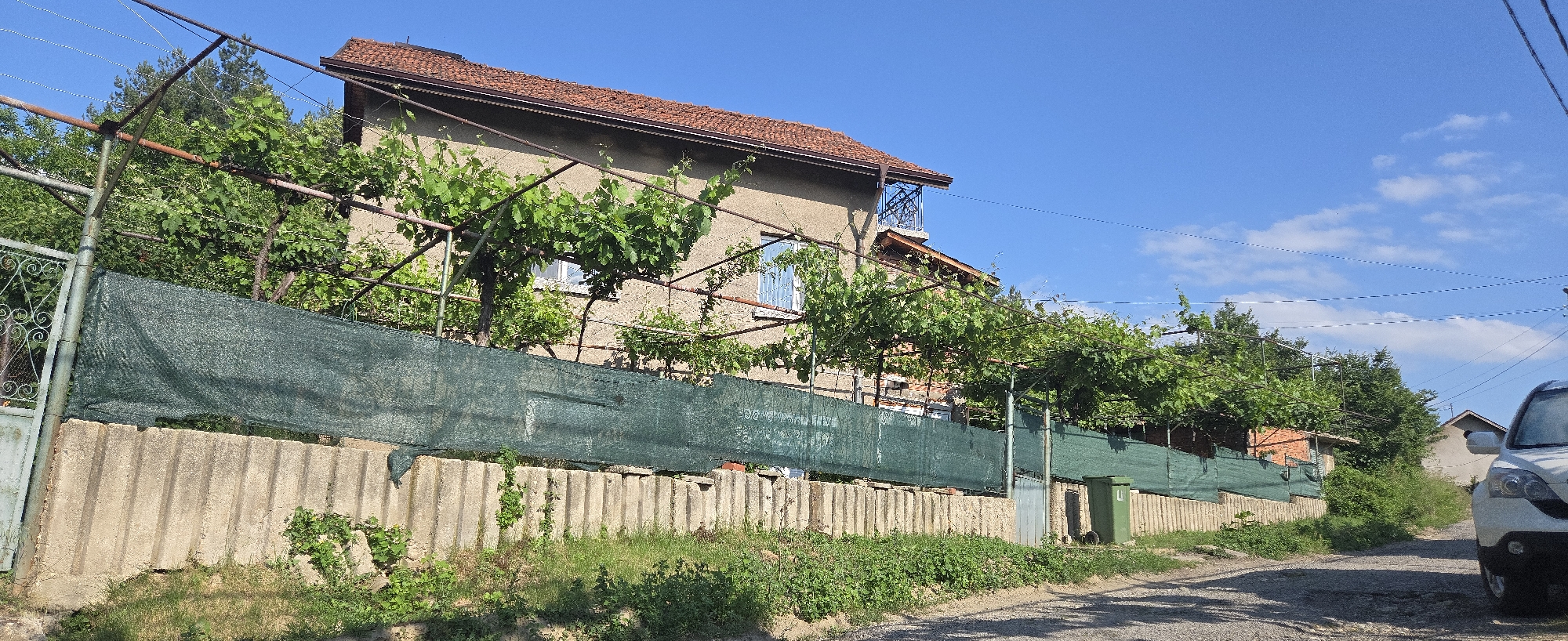 This image depicts a residential street scene under a clear blue sky with sparse clouds. In the foreground, an unpaved road with a gravel and dirt surface shows shadows from adjacent structures. To the left, a concrete block wall serves as the base of a fence, topped by a dark green mesh fabric acting as a privacy screen. A metal gate with decorative scrollwork is visible on the far left of this fence. Behind the fence, a robust metal framework supports a dense canopy of green grapevines or similar foliage, creating an overhead arbor.
Behind the vine-covered structure stands a two-story house with a reddish-brown tiled roof and light brown or beige stucco-like exterior walls. The upper floor features at least two windows; one on the left appears to have a protective metal grill, and another further right has white, possibly louvered, coverings. A small balcony with an ornate black metal railing projects from the upper right side of the house. Utility lines are visible strung across the upper right portion of the sky.
Further along the fence to the right, a dark green waste bin is positioned near the roadside. On the far right, the front-right section of a white SUV, including its headlight and bumper, is partially visible, suggesting it is parked or moving on the road. Lush green vegetation, including grass, weeds, and distant trees, is present throughout the scene. No individuals are visible. The location is in the city of Unknown, Unknown.