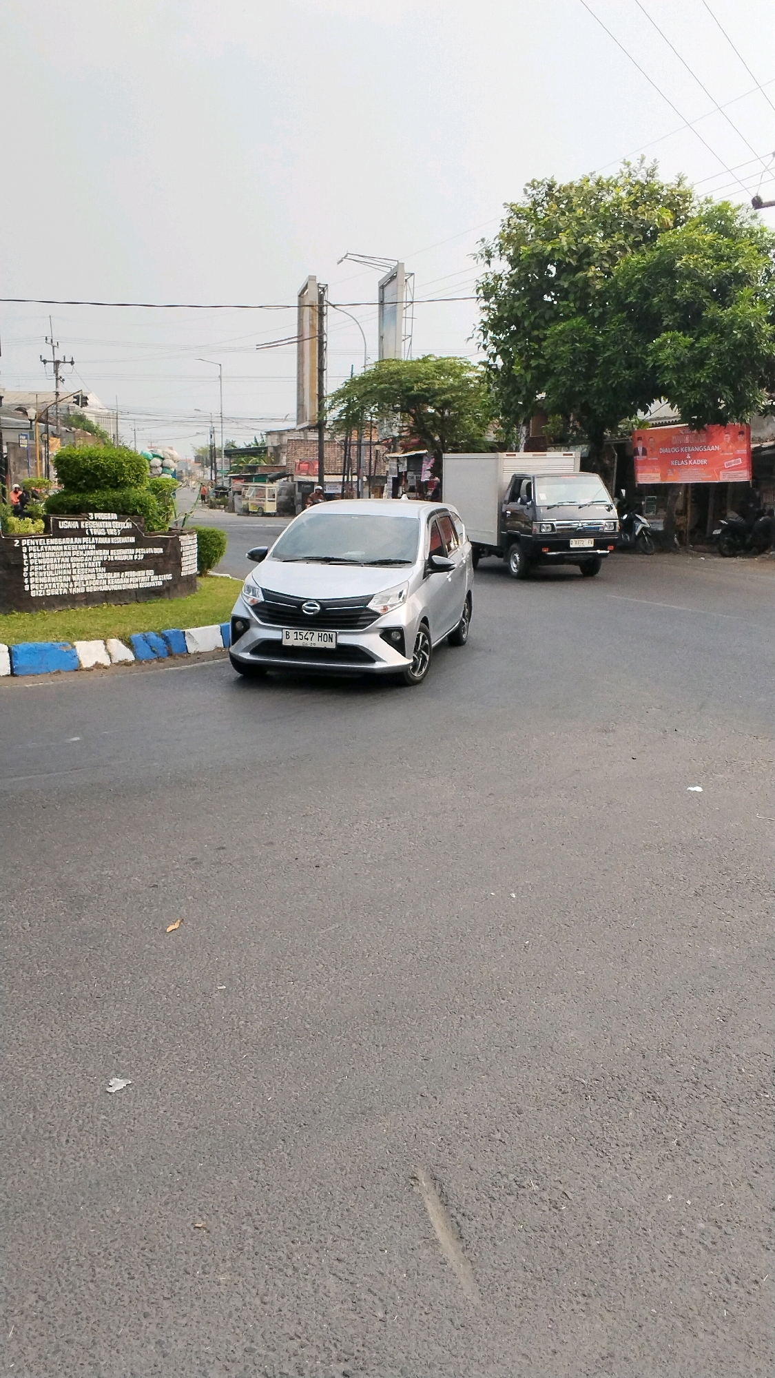 The image depicts a street scene in Banjaragung, Indonesia, under an overcast sky. A silver Toyota Calya hatchback, license plate B 3567 HON, is prominently positioned in the foreground, driving on an asphalt road. Behind it, a white box truck, possibly a light commercial vehicle, is also on the road, followed by a black pickup truck with license plate D 9372 FX.
To the left of the road, a monument with text inscribed on it, surrounded by a manicured hedge and a curb decorated with blue and white stripes, stands. Several street lights and power lines crisscross the sky. A large, leafy green tree is visible on the right side of the image, partially obscuring a banner with red text that reads "DIALOG KERABANGSAAN KELAS KADER". In the background, various buildings, including what appear to be shops and houses, line the street, along with more trees and utility poles. The overall environment suggests a mixed urban and residential area. The asphalt road shows some minor wear and tear, with a few small pieces of debris scattered on its surface.