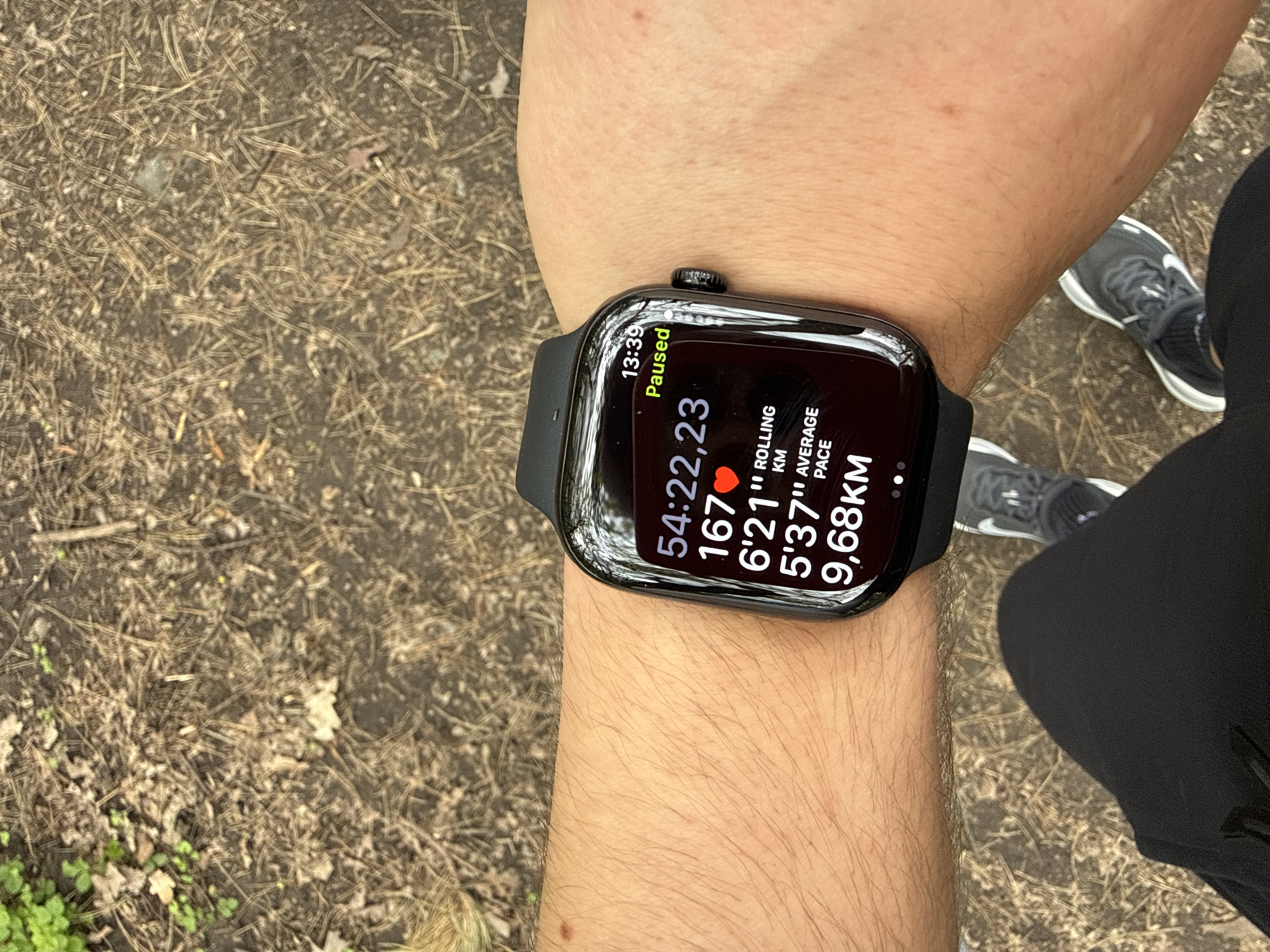 A close-up, high-angle shot shows a person's arm wearing an Apple Watch, with the watch face displaying running data: a time of 54 minutes, 22 seconds, 23 milliseconds, a heart rate of 167 beats per minute, a rolling kilometer time of 6 minutes, 21 seconds, an average pace of 5 minutes, 37 seconds, and a distance of 9.68 kilometers. The watch face also indicates that the workout is "Paused" and the time is 13:39.
The person is wearing black running tights and Nike running shoes, partially visible in the background. The surrounding environment consists of an outdoor path or trail covered in fallen leaves and pine needles, and scattered greenery.
The location is likely somewhere in Sofia, Bulgaria.