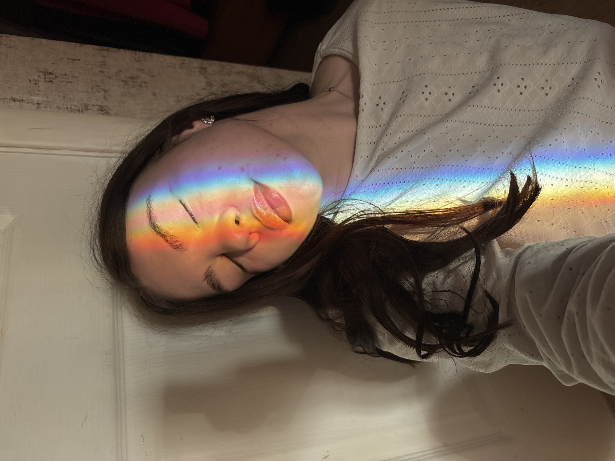 A woman is lying down, angled toward the viewer. Her face is illuminated by a rainbow light. She has dark hair, and is wearing a white, eyelet-style top. The background is a neutral color. The location is unknown; there is no indication it is in Perm, Russia.
