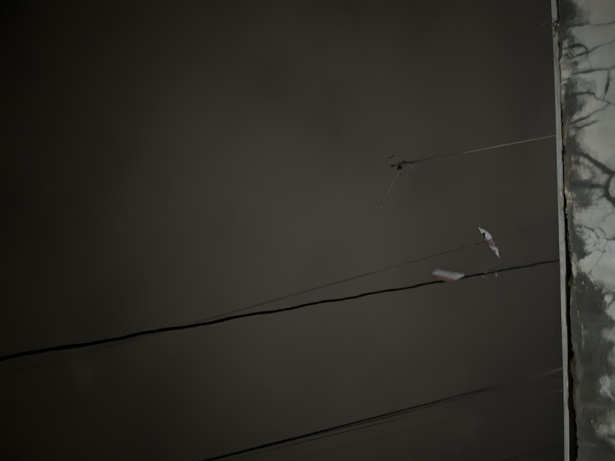 The image shows a dark, overcast sky with several power lines running diagonally across the frame. A weathered concrete wall is visible at the bottom of the image. A TV antenna is positioned in the upper right quadrant. Two small, colorful objects, possibly paper kites or flags, are suspended from one of the power lines. The overall scene suggests an outdoor environment in a somewhat urban or suburban setting, possibly during twilight or a gloomy day. The specific location in Pekalongan, Indonesia cannot be determined from the visual information alone.
