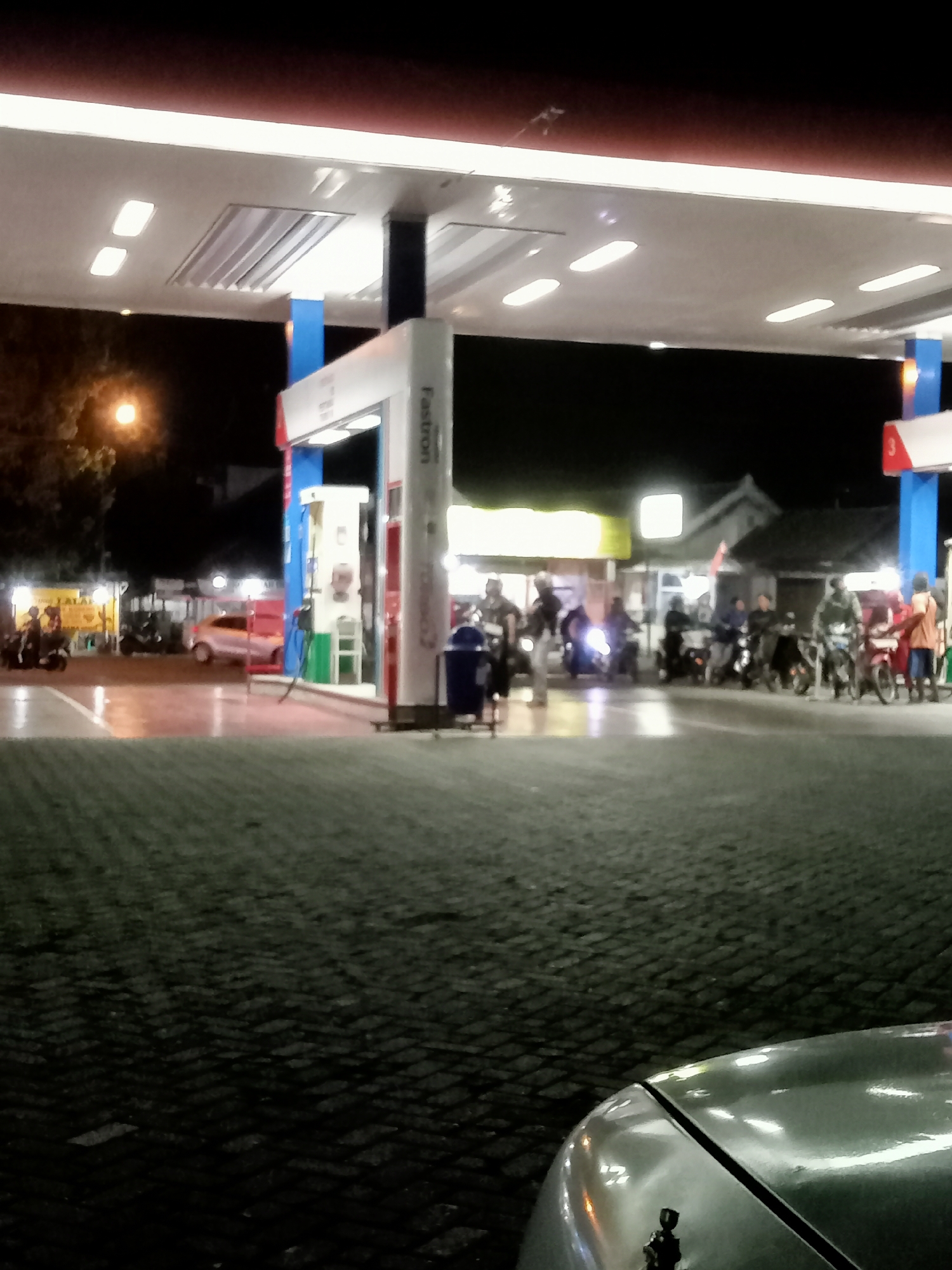 The image depicts a night scene at a Fastron gas station in Plosokandang, Indonesia. The gas station is illuminated by overhead lights, casting a glow on the wet, cobblestone-like ground. Several motorcycles and a car are parked or moving around the pumps. People are visible, some on motorcycles and others standing near the pumps. A gas pump with the "Fastron" logo is prominent in the foreground. The overall atmosphere suggests a busy, late-night refueling stop.