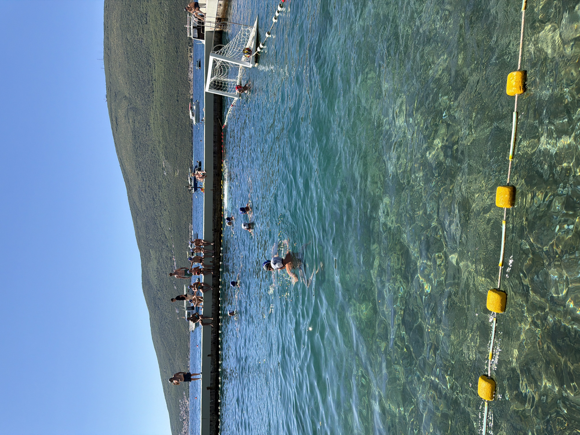 In Đenovići, Montenegro, a bright sunny day is depicted at an outdoor swimming area. Several people are gathered on a concrete pier that borders the clear, turquoise water. Some are sitting on the edge, while others stand. In the water, a group of individuals, many wearing blue caps, are engaged in what appears to be a water polo game, with a goal visible on the right side of the frame. The water is shallow enough in the foreground to see the rocky seabed. A line of yellow buoys with a rope separates the swimming area. In the background, green, tree-covered hills rise, and several boats are anchored in the bay under a clear blue sky.