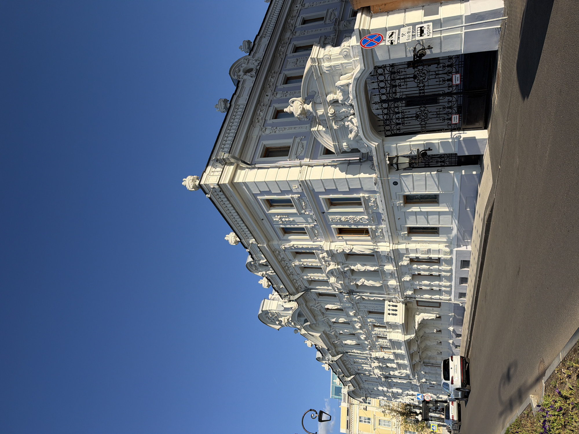The image captures a wide, low-angle view of a grand, white, ornate building facade on a sunny day with a clear blue sky. The building features intricate stucco decorations, including sculptures of human figures and elaborate patterns, along its multiple stories and roofline. A large, black wrought-iron gate is visible on the right side of the building, adorned with sculptural elements above it. A street runs in front of the building, with two cars parked on the side and one driving away. Traffic signs are present near the gate, one indicating no stopping and another with time restrictions and a symbol for buses. A vintage-style lamppost stands on the left side of the street, casting a shadow on the asphalt. In the background, a yellow building with multiple windows can be seen. This building is identified as the former mansion of the merchant G. A. Gubin in Nizhny Novgorod, Russia.