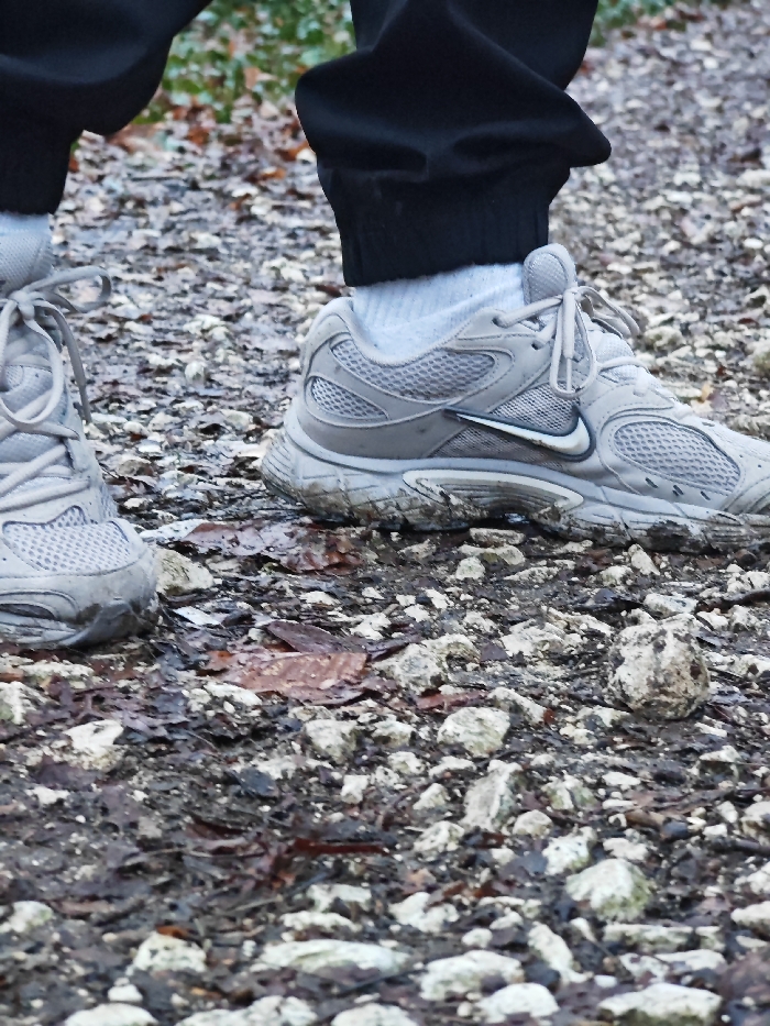 The image presents a low-angle, close-up view of an individual's lower legs and feet on an unpaved, uneven surface in Shumen, Bulgaria. The terrain is composed of numerous small, light-colored stones, dark brown soil, and scattered wet, decaying brown leaves. The person is wearing black trousers with elasticated cuffs, light grey/white socks, and light grey athletic shoes. The right shoe, featuring a visible Nike 'swoosh' logo on its lateral side, and the left shoe both exhibit accumulated mud and debris on their soles and lower sides, consistent with outdoor traversal. Blurred green and brown foliage is discernible in the upper background, indicating a natural or semi-natural outdoor environment, potentially a park or trail within the city.
