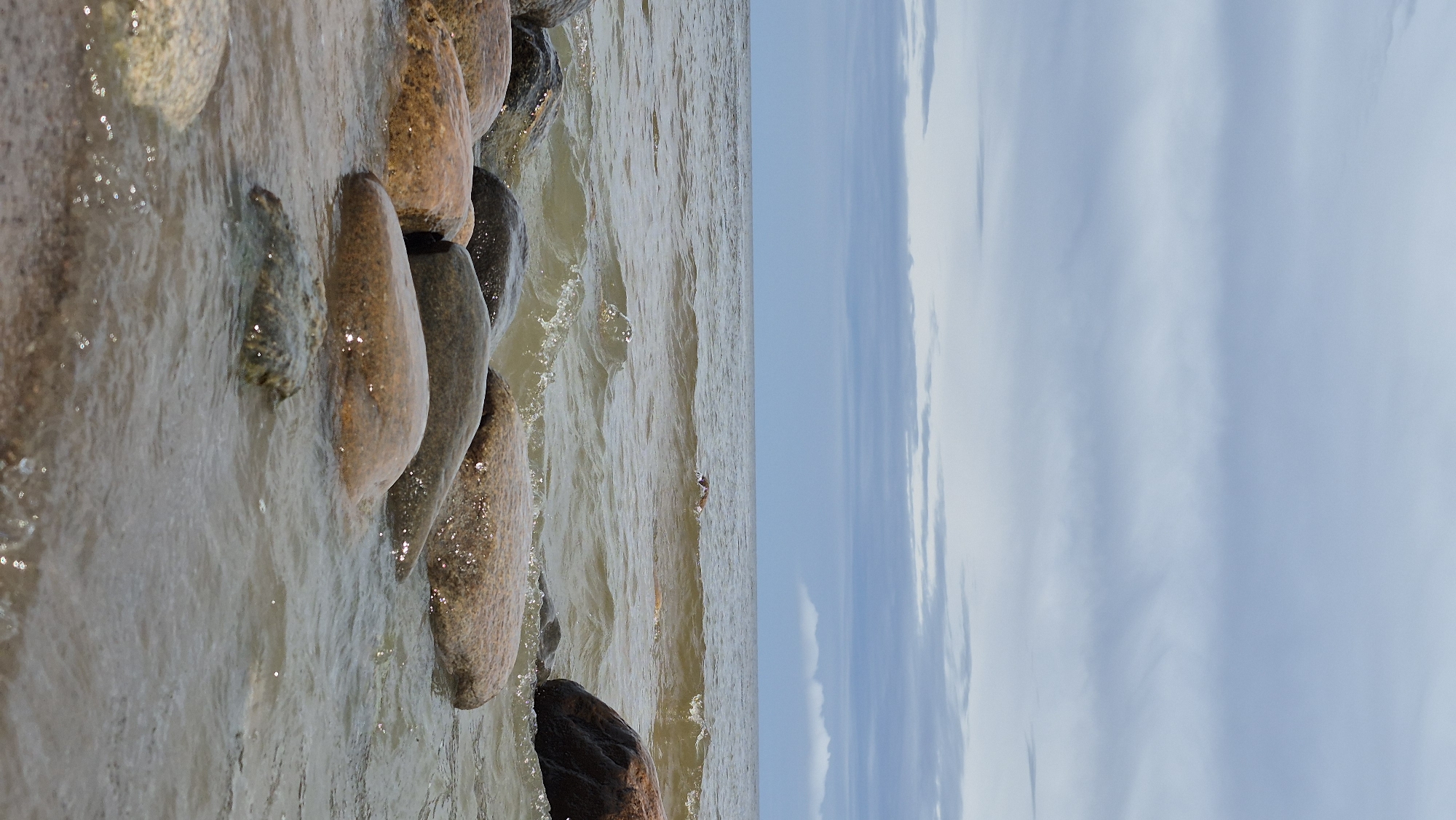 The image depicts a rocky shoreline on a cloudy day. Large, smooth stones of various earth tones are partially submerged in the gently lapping water. The sea is a muted brown-green, reflecting the overcast sky. The sky itself is filled with layered, horizontal clouds, varying in shade from light grey to a deeper blue-grey. The composition is dominated by the natural elements of water and stone, with no human presence visible.