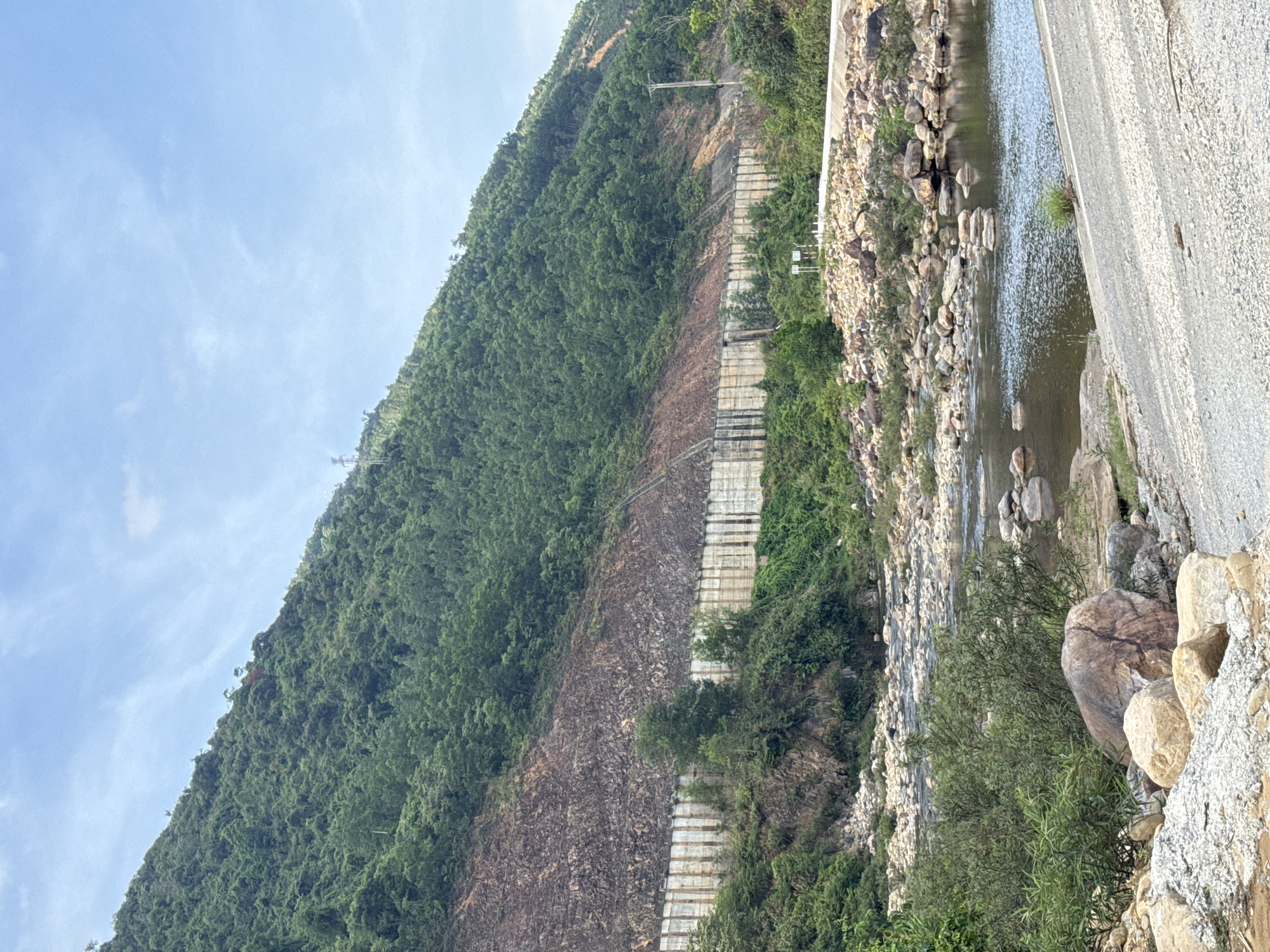 The image depicts a landscape likely in Đà Nẵng, Vietnam. A heavily vegetated mountain slopes downward to a river. Along the slope, a terraced retaining wall structure is visible, possibly for erosion control. To the right of the image is a concrete barrier or roadway that appears to edge along the river's bank. Boulders are scattered along the river's edge. The sky is partially obscured by the mountain but shows a light blue color with some cloud cover.