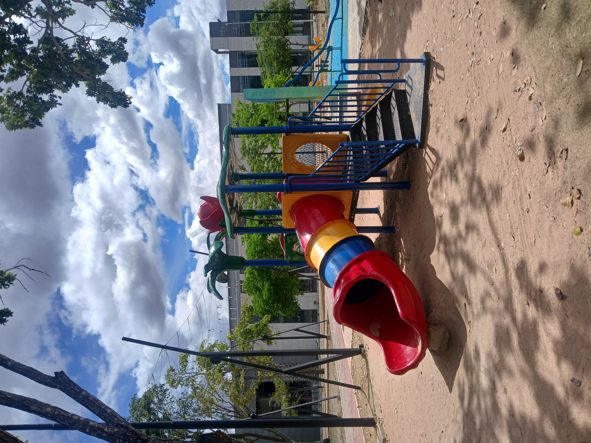 The image shows a colorful playground slide in a park in Ciudad Guayana, Venezuela. The slide has red, yellow, and blue segments, with a red exit. The slide is connected to a structure with blue railings and black stairs. The ground is a mix of sand and dirt, with scattered leaves. The sky is blue with large white clouds. Trees and buildings are visible in the background. There are no people in the immediate vicinity of the slide. The image is taken at an angle, making the playground appear tilted. A power pole with wires is in the background on the left.