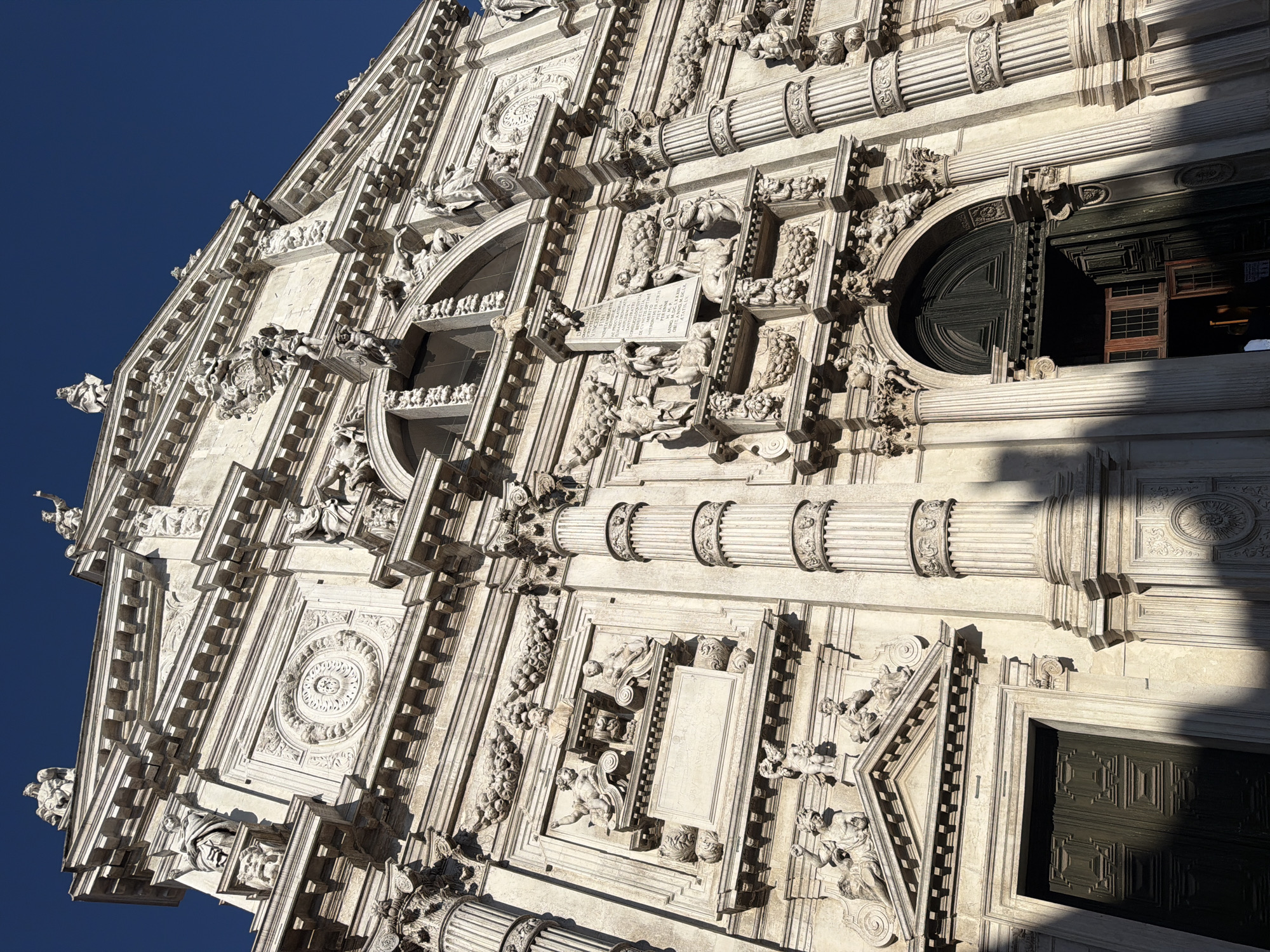 The image shows a low-angle, full-frame shot of the facade of a large, ornate building, likely a church, under a clear blue sky. The architecture is predominantly white or light-colored stone and features intricate sculptural details, including statues, reliefs, and decorative moldings. The facade is symmetrical with a grand central entrance and two smaller entrances on either side. Columns, both fluted and engaged, support pediments and entablatures. The upper section of the facade is crowned with statues and decorative elements. Sunlight casts strong shadows, highlighting the depth and texture of the carvings. The style of the architecture suggests a Baroque or Renaissance influence. Based on the distinctive architectural style and ornate decoration, this is the facade of the Basilica dei Santi Giovanni e Paolo (San Zanipolo) in Venice, Italy. There are no people visible in the image.
