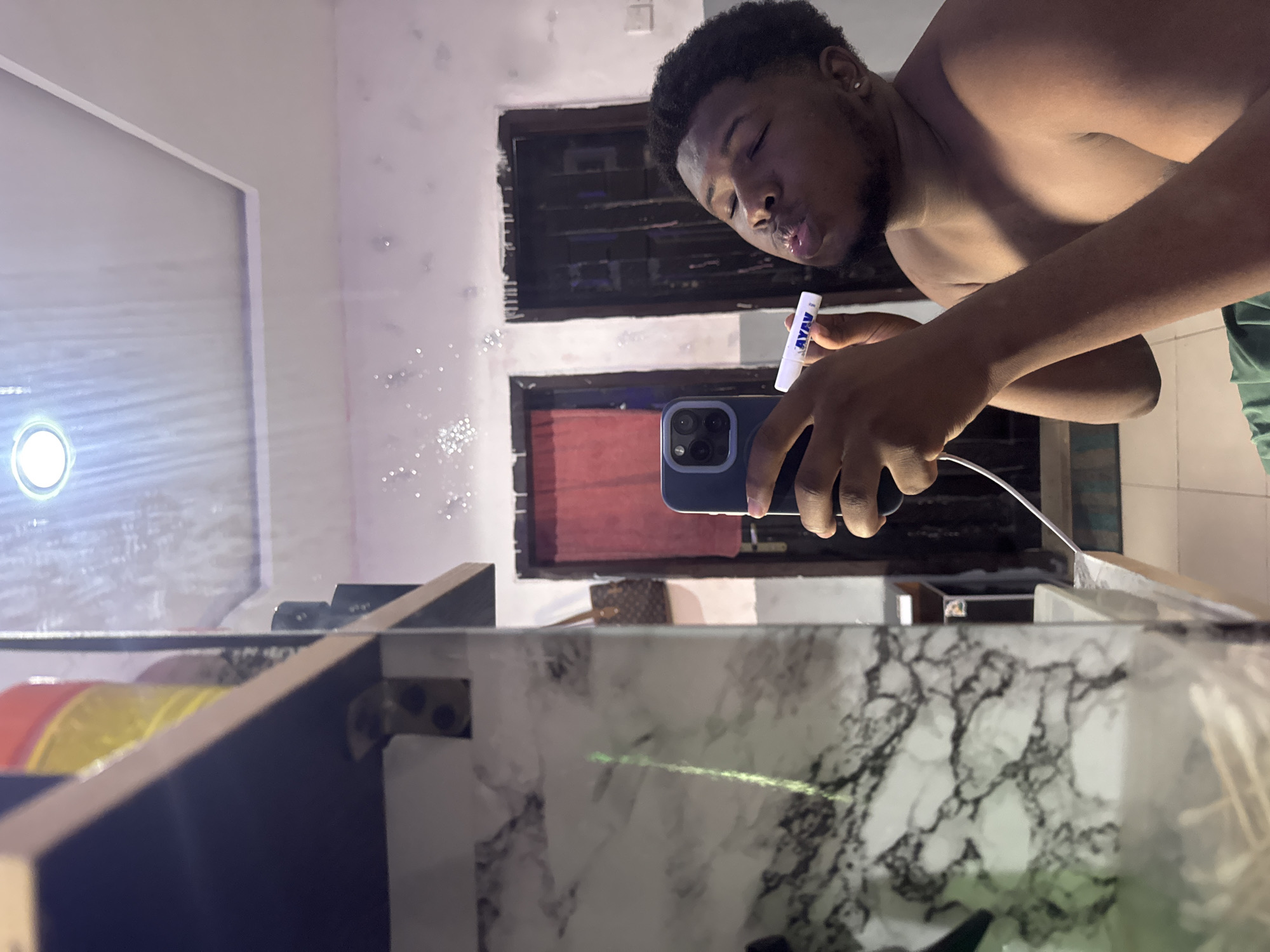 A shirtless Black male in Gbongan, Nigeria holds a blue mobile phone in his left hand while blowing bubbles with a white bubble wand held in his right. He is likely taking a selfie in front of a bathroom mirror. The bathroom has dark wooden shelves and a marble countertop. A small white light is fixed above the mirror. Two dark wooden windows with visible shutters sit behind the man's reflection in the mirror.