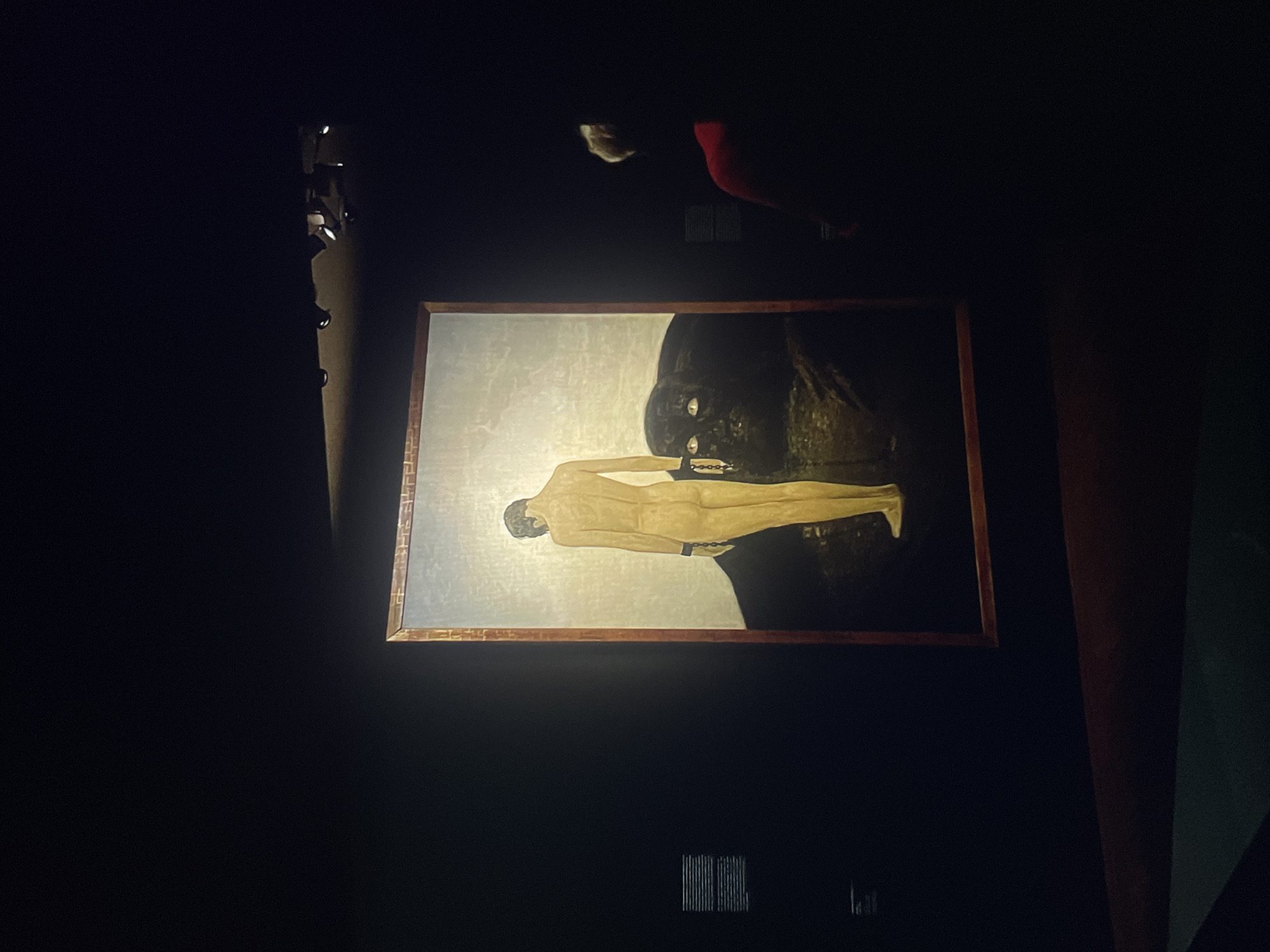 The image shows a framed painting in a dimly lit room, possibly a gallery or museum. The painting depicts a nude male figure with chains on his wrists and ankles. The figure is lying horizontally against a dark, indistinct background that might include another figure or object. A person with white hair and wearing a red garment is visible in the background. To the left of the painting are several light fixtures.
Location: Unknown, Unknown.