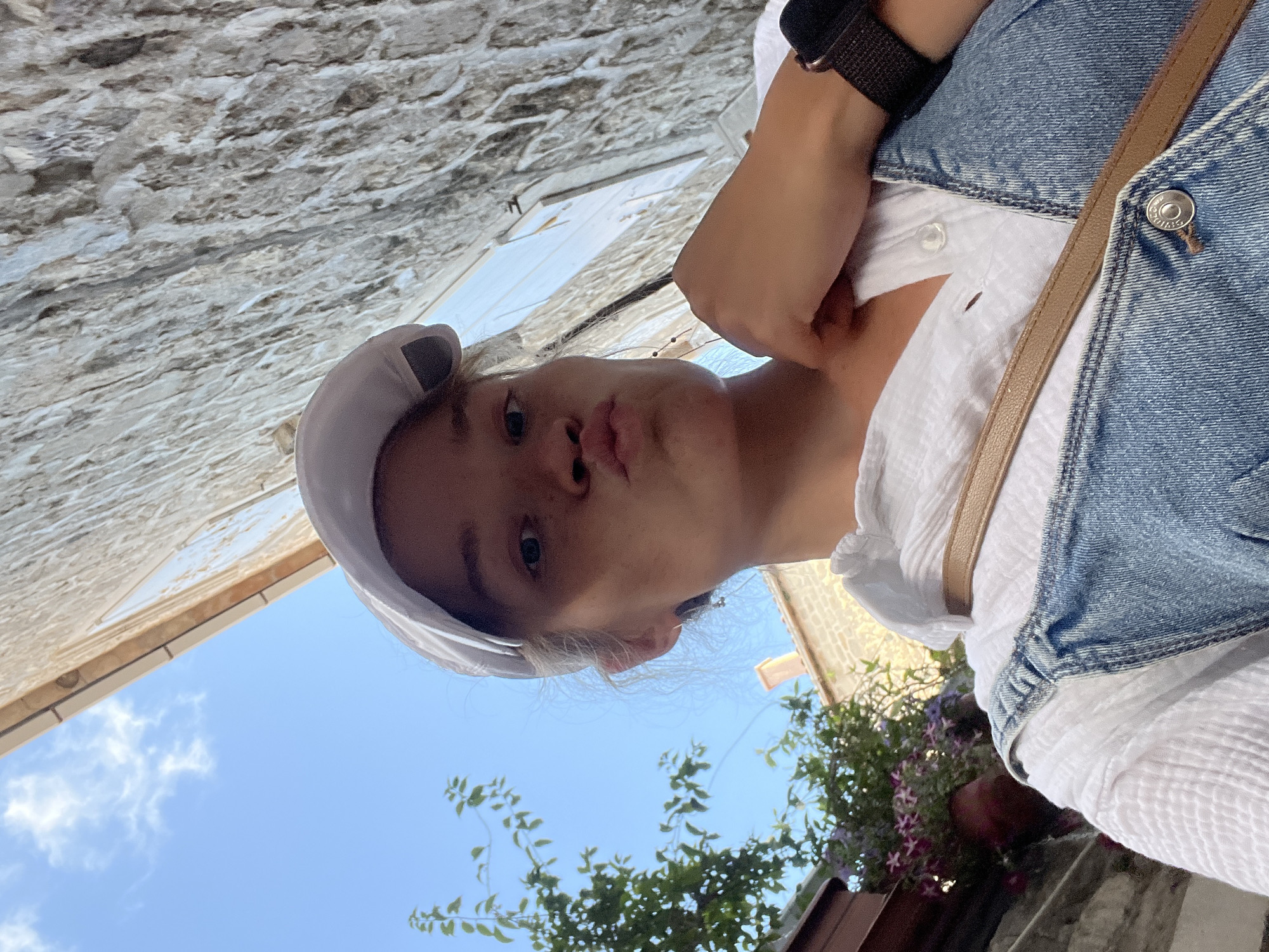 A young person, appearing female with light hair, is captured in a low-angle selfie, looking up towards the camera. She has a playful expression with puckered lips. She wears a light-colored headband or cap, a textured white shirt or top, and blue denim overalls or a pinafore dress. A brown leather strap from a bag crosses her chest, and a dark watch or wristband is visible on her left arm.
The background features a rough, light-colored stone or stucco wall, indicating an older architectural style. Above and to the right, a clear blue sky with a few faint white clouds is visible, suggesting bright daylight. In the lower right corner, lush green foliage with small clusters of purple flowers, likely in a planter or hanging basket, adds a touch of natural beauty. The overall scene conveys a sunny outdoor setting, consistent with a Mediterranean town like Budva, Montenegro. A small, silver button on the denim garment has an embossed design or illegible text.