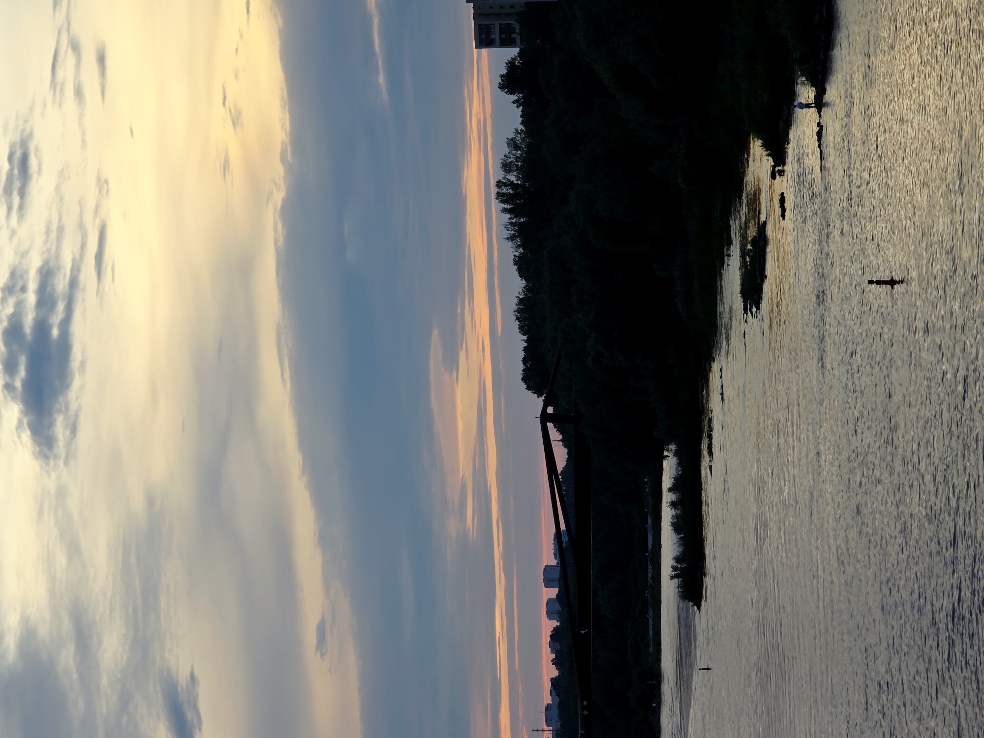 The image depicts a river scene at dusk in Warsaw, Poland. The sky is filled with a gradient of colors, transitioning from pale yellow at the top to pink and blue hues closer to the horizon, with scattered clouds. A dark silhouette of trees lines the far bank of the river, with a bridge structure visible in the midground. Beyond the trees, several buildings and a tall apartment building are silhouetted against the sky. The river itself reflects the sky's colors, with rippling water creating a textured surface. In the foreground of the river, a dark marker or buoy is visible. Further along the bank, a solitary figure, silhouetted and wearing blue, stands on some rocks near the water's edge, possibly fishing or observing. Another person is visible as a dark silhouette further to the left, near the bank. The overall atmosphere is tranquil and serene.