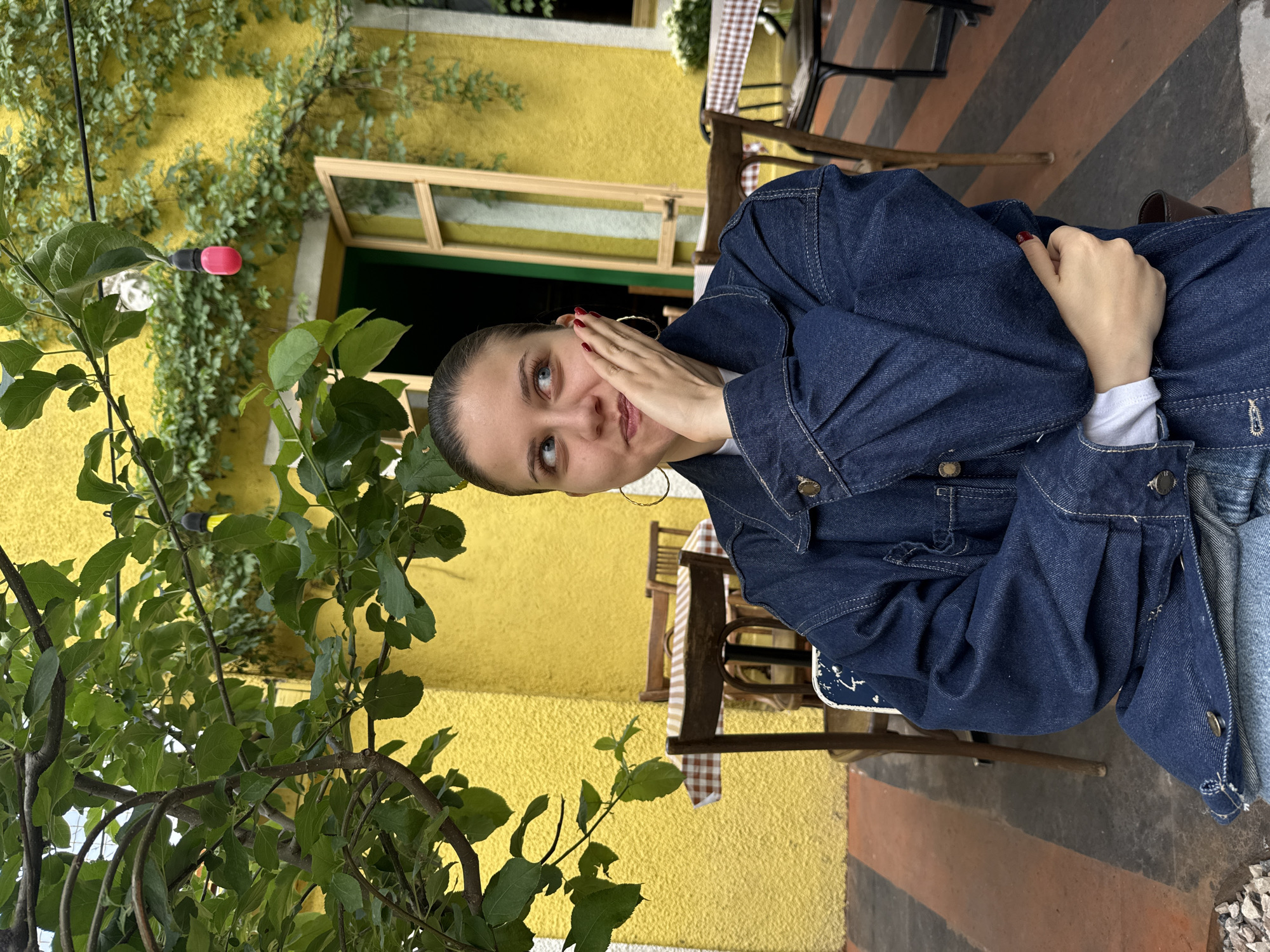 A slightly rotated medium shot shows a young woman seated in an outdoor setting, seemingly a cafe or restaurant patio. She is wearing a dark denim jacket over a white top and large hoop earrings, with her right hand resting on her face. The woman is looking upwards and to her left with a pensive or thoughtful expression.
Behind her, there's a bright yellow building with an open window framed in light wood. The building is partially covered with climbing foliage. A table covered with a brown and white checkered tablecloth and chairs are visible in the background, indicating a seating area. The ground has alternating stripes of gray and a rusty orange color.
On the left side of the frame, there's a tree with green leaves and a visible pink decorative light. The location can be inferred to be outdoors, possibly in a public space such as a restaurant patio or garden. The city of Yerevan, Armenia is not obviously evident from this image alone.