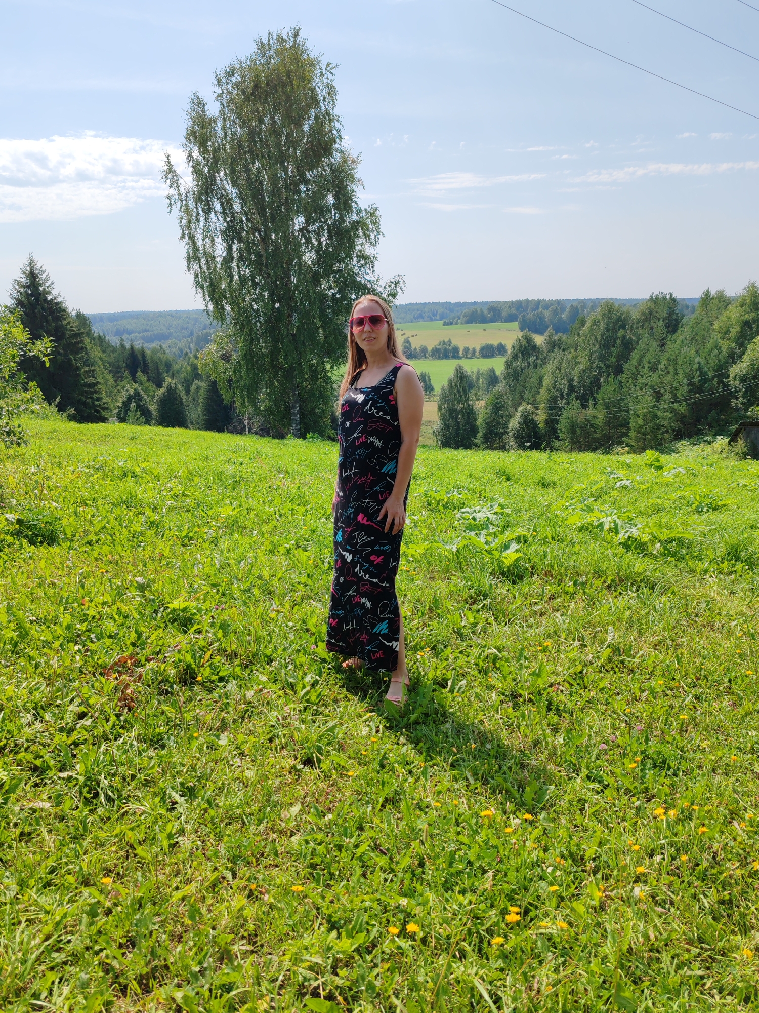 In Чухлэм, Russia, a young woman stands in a sun-drenched grassy field. She is wearing a long, black, sleeveless dress with a colorful pattern of scribbled words and designs. She has on pink sunglasses and her hair is light brown. The field is covered in various green grasses and small yellow wildflowers. In the background, rolling hills covered with dense green trees stretch towards the horizon under a clear blue sky with scattered white clouds. A tall, slender birch tree stands to the left of the woman, and a few other trees are visible closer to the foreground on the left. Power lines are visible in the upper right of the image.