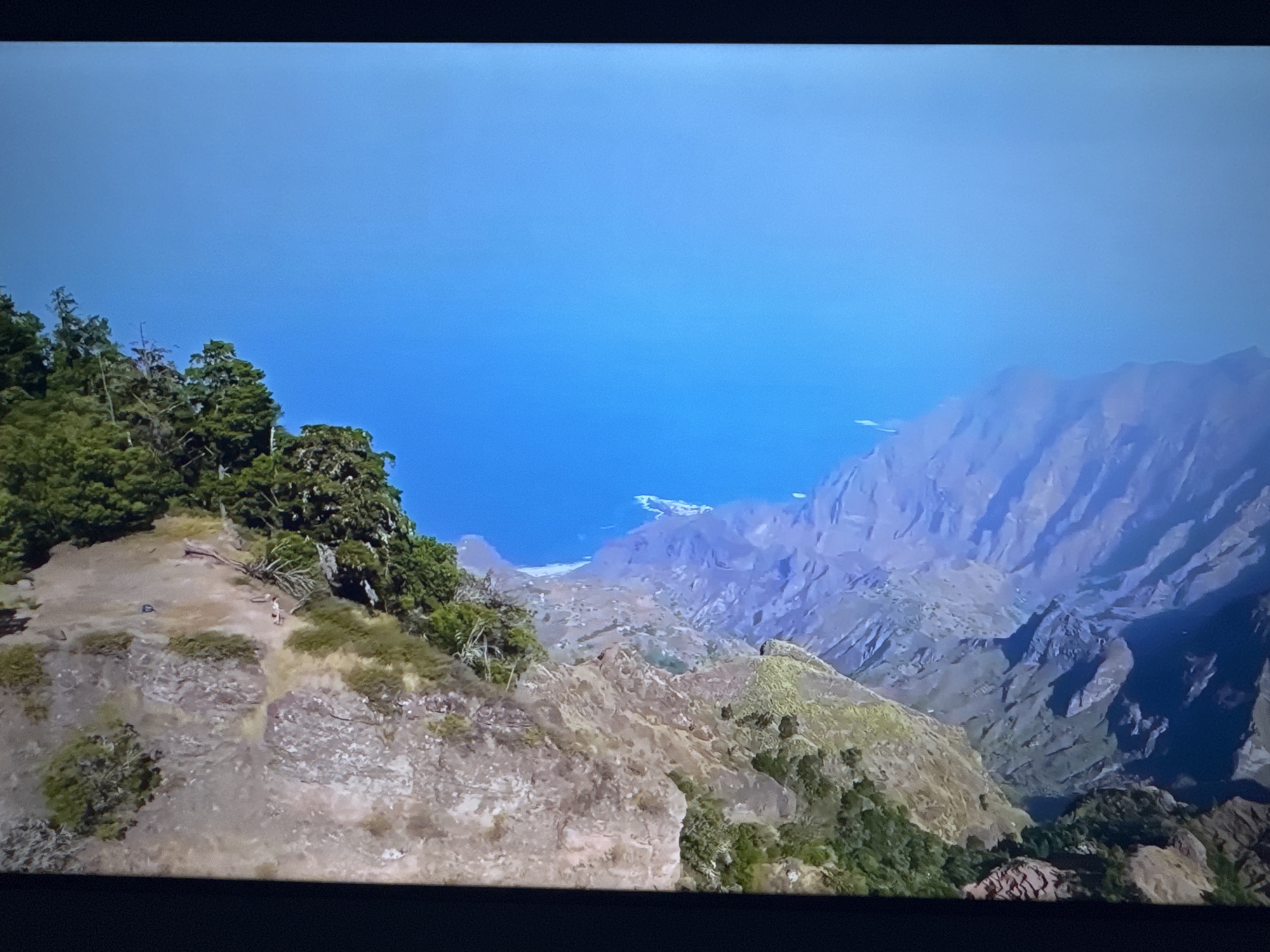 The image shows a panoramic view of a rugged, mountainous coastline under a clear blue sky. In the foreground, a steep, rocky slope is covered with sparse vegetation and a few trees. A small figure, possibly a hiker, can be seen on a dirt path traversing the slope. Further in the distance, the terrain descends into deep valleys and ridges, revealing layers of rock formations that lead to the sea. The ocean is a vibrant blue, meeting the land at a hazy horizon. There is no indication of the city of Ləcət, Azerbaijan, in this image.