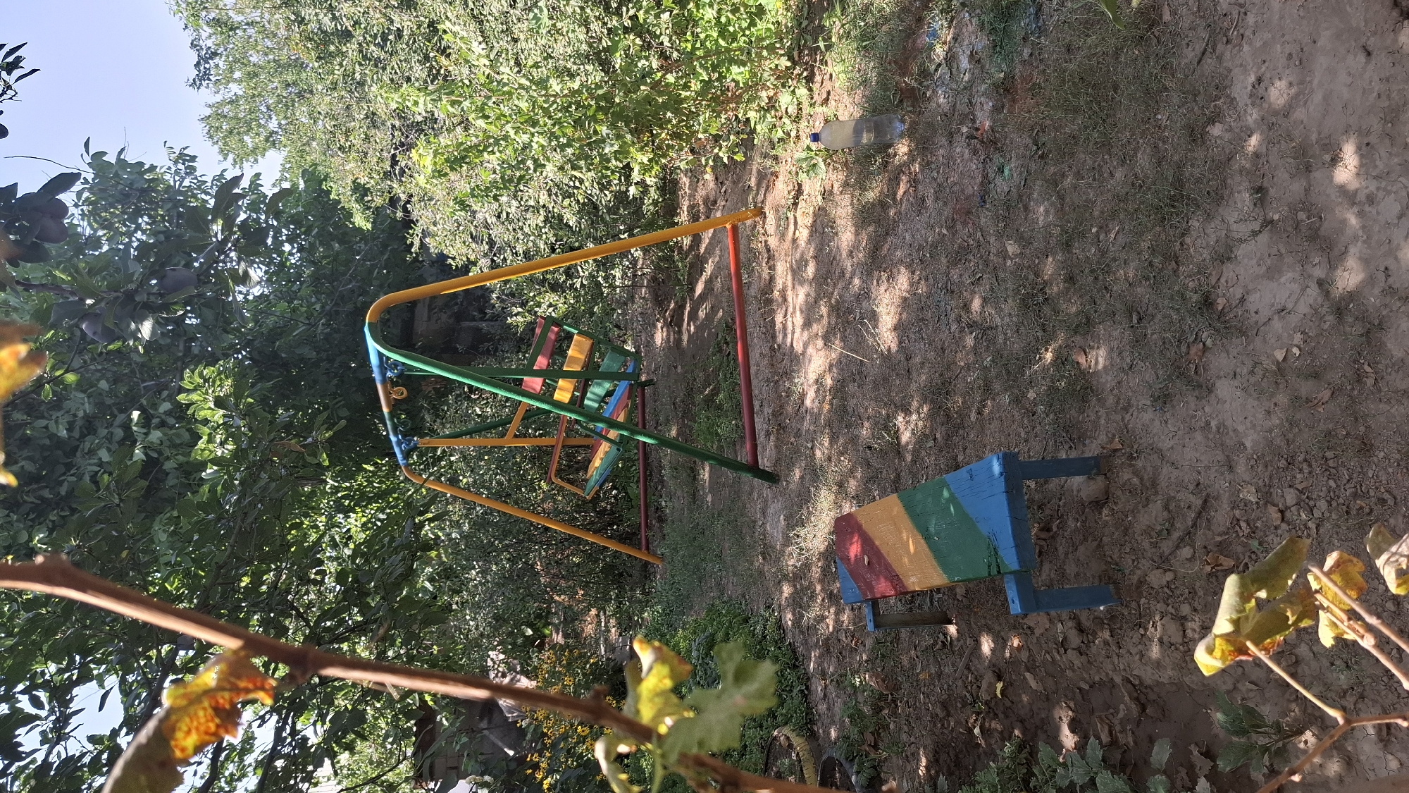 The image shows an outdoor scene in the city of Shubarssu, Kazakhstan. A brightly colored children's swing set with a yellow, green, and blue frame and a multi-colored seat is the central focus. In front of the swing set, a wooden bench, also painted with multi-colored stripes of red, yellow, green, and blue, sits on the dirt ground. The ground is uneven and covered with dry grass and fallen leaves. Lush green trees and foliage surround the area, with some branches and leaves in the foreground, suggesting a garden or park setting. A plastic water bottle is visible on the right side of the image, near the foliage. The sky is visible in the upper portion of the image, with a clear blue sky. The overall impression is a simple, possibly homemade, play area in a natural, somewhat unkempt, environment. There are no people present in the image.